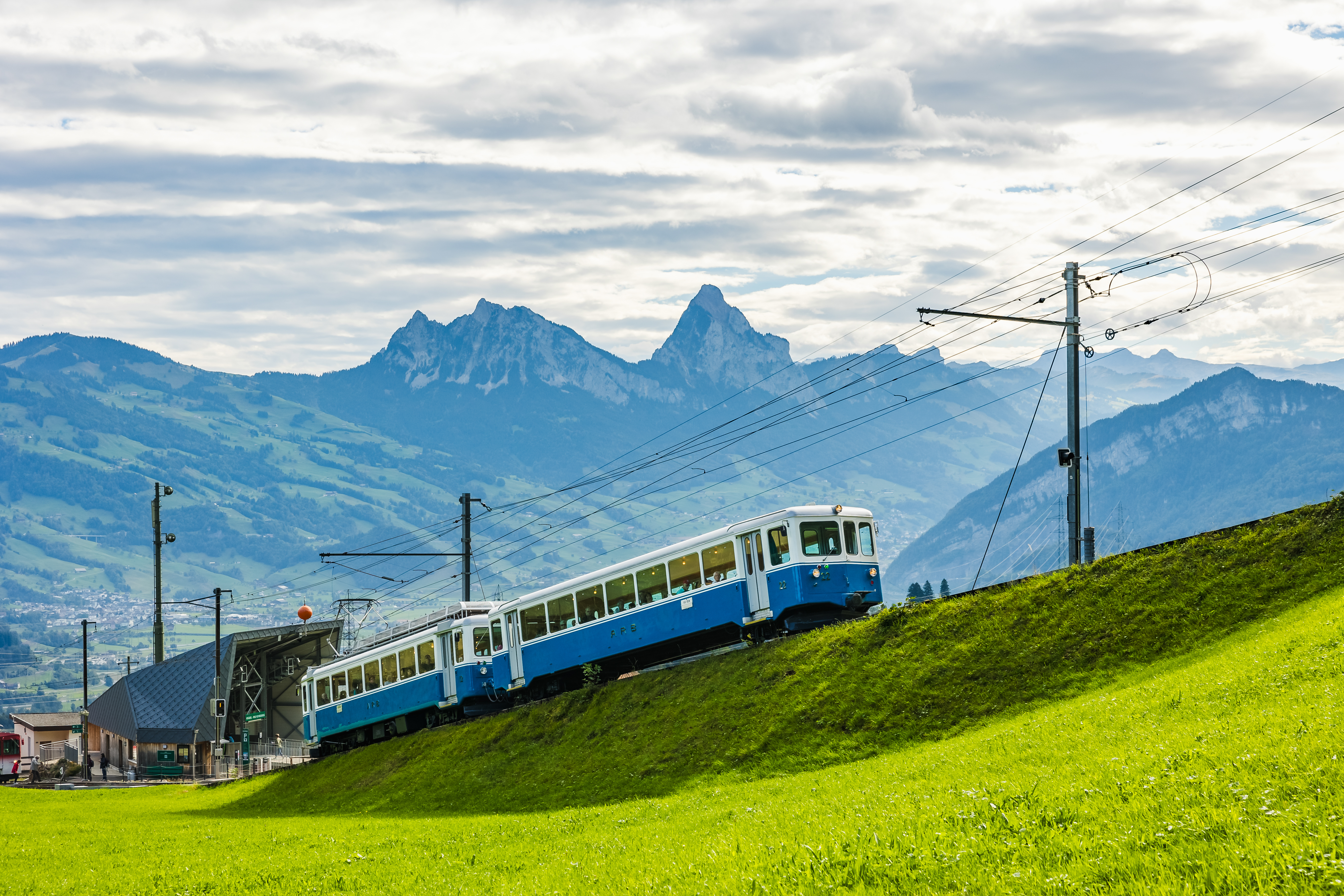 Blauer Nostalgiezug der Rigi Bahnen fährt bei der Station Kräbel mit Blick auf die Mythen durch die grüne Zentralschweizer Berglandschaft.