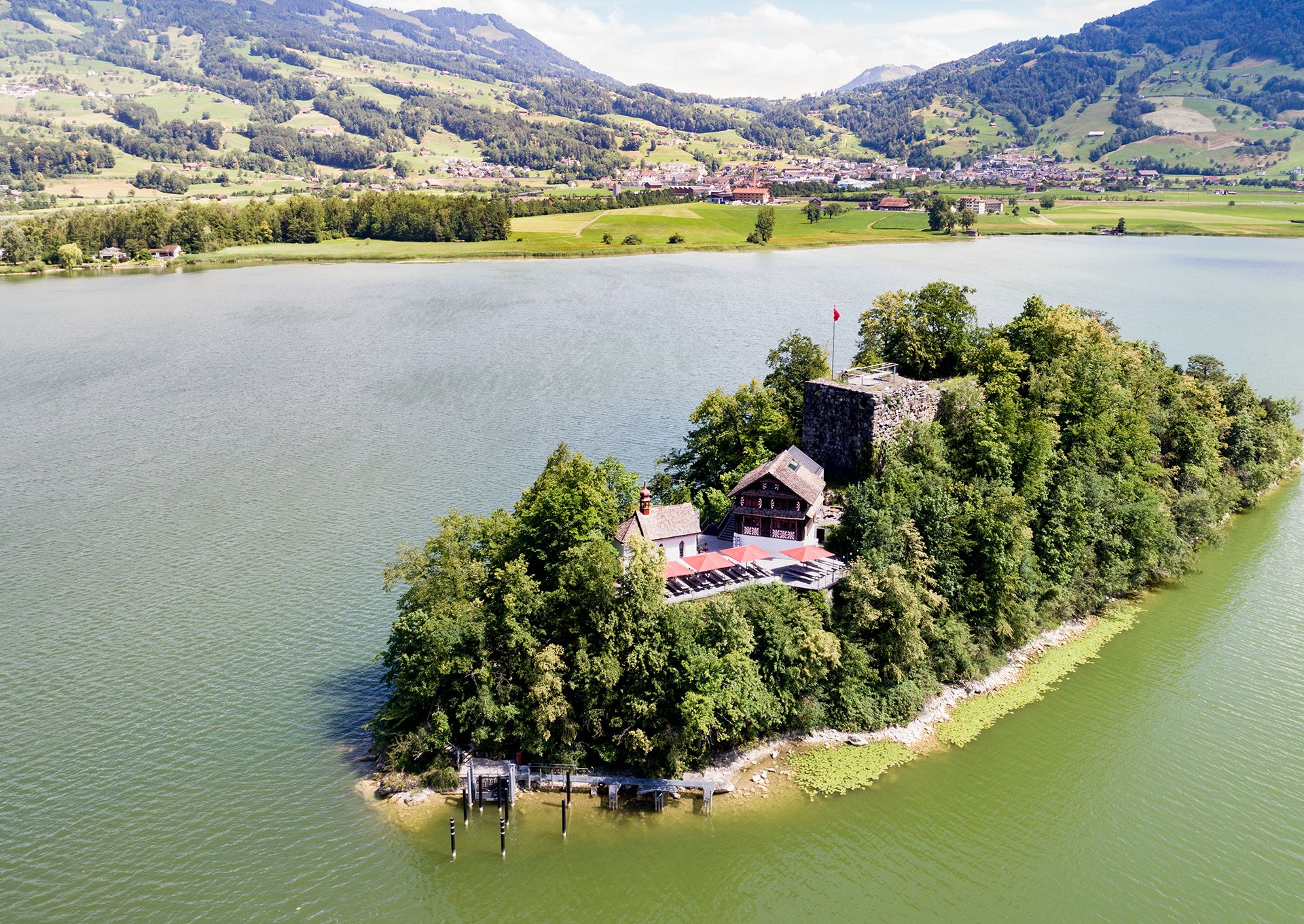 Vue aérienne de l’île de Schwanau sur le lac de Lauerz, avec son auberge et sa ruine historique, entourée d’eau verte et de collines.
