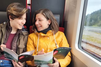 Zwei Frauen sitzen im Zug und blättern in einer Broschüre