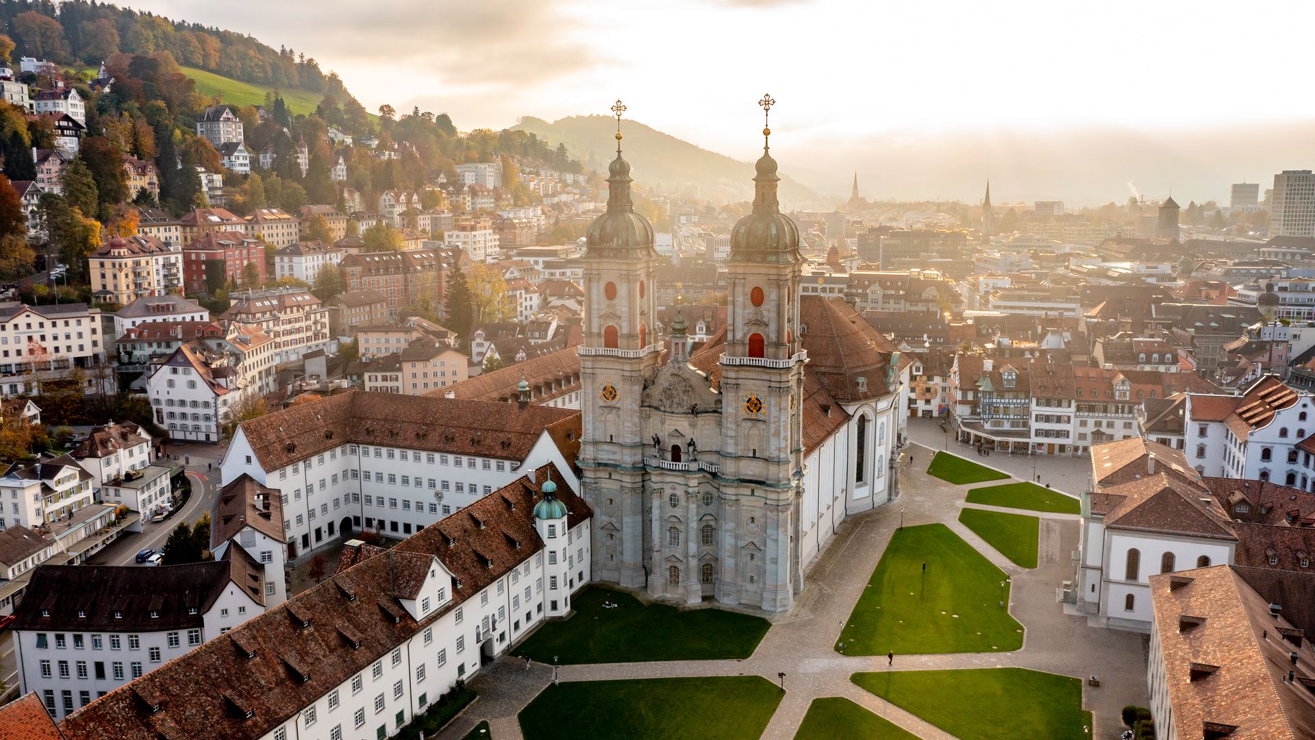 Aerial view of the St. Gallen monastery district and cathedral, surrounded by historic buildings and green lawns, with the picturesque town in the background and the hills in the foreground.