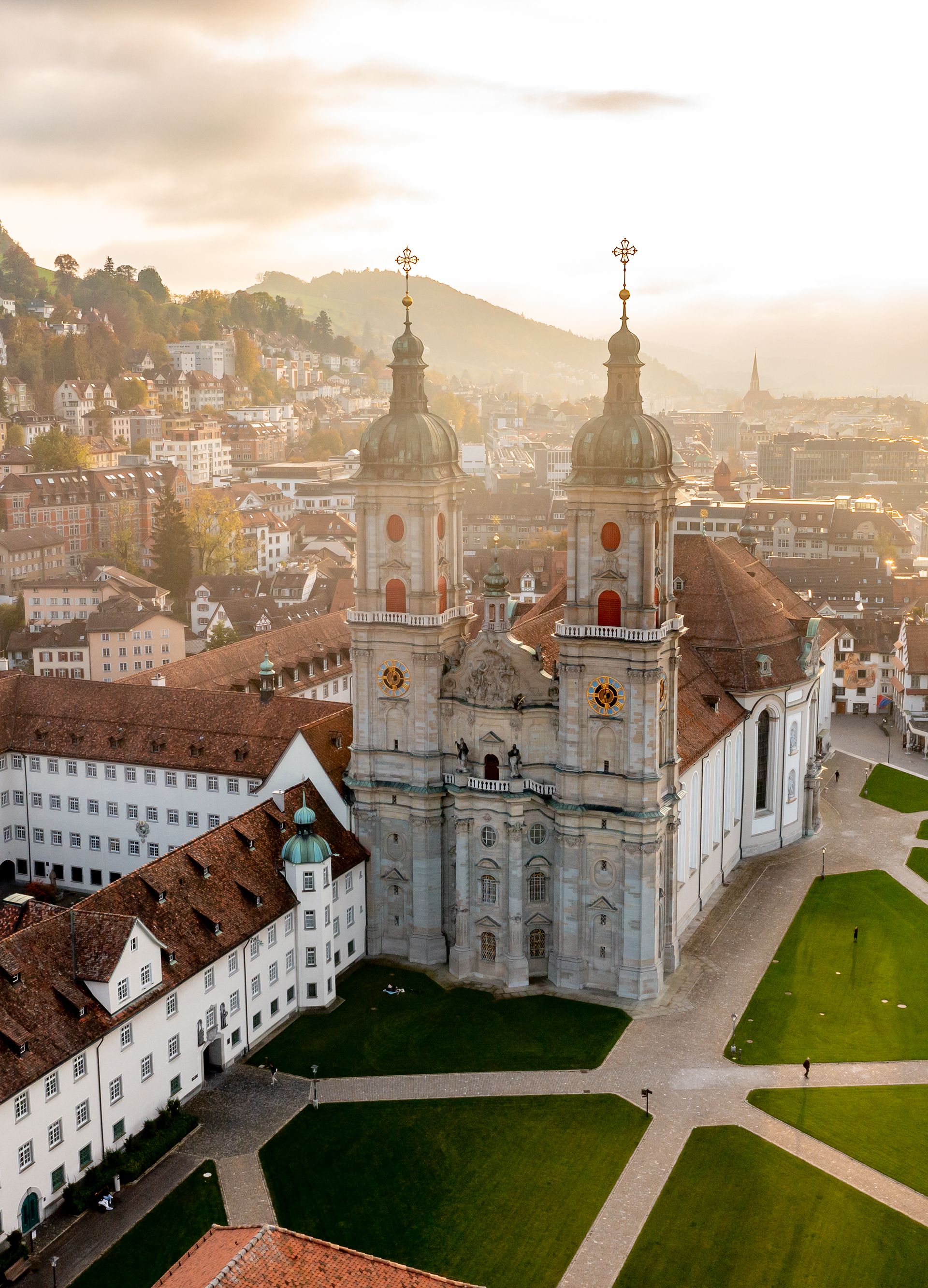 Aerial view of the St. Gallen monastery district and cathedral, surrounded by historic buildings and green lawns, with the picturesque town in the background and the hills in the foreground.