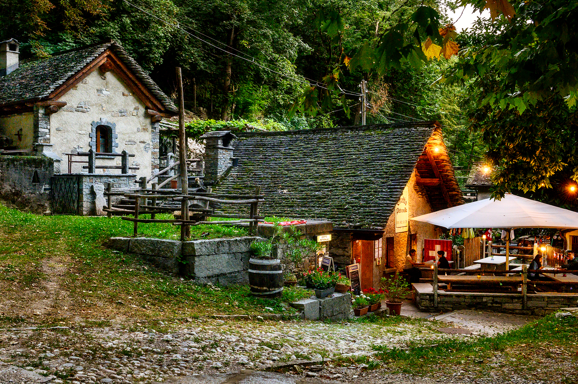 Photo d'ambiance prise en soirée du Grotto Petronilla à Biasca, avec ses maisons traditionnelles en pierre, sa terrasse éclairée et son atmosphère chaleureuse dans la verdure.