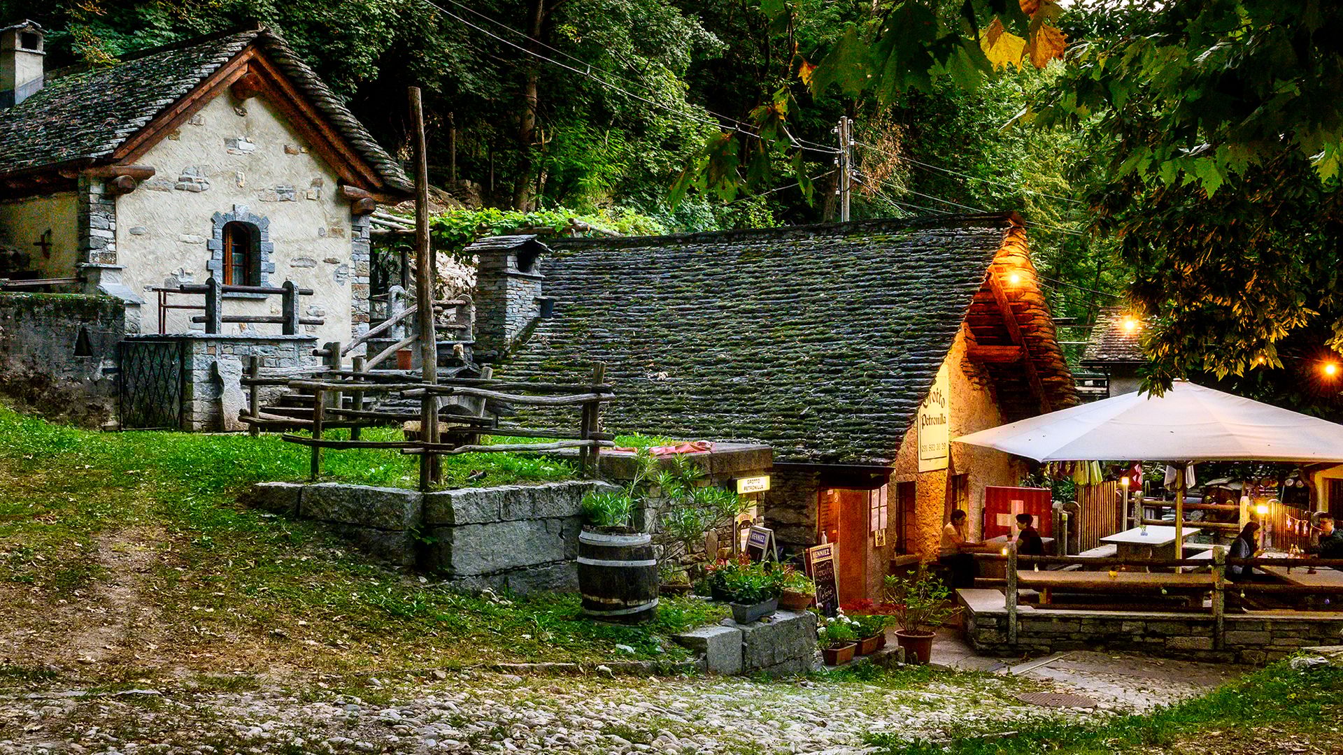 Photo d'ambiance prise en soirée du Grotto Petronilla à Biasca, avec ses maisons traditionnelles en pierre, sa terrasse éclairée et son atmosphère chaleureuse dans la verdure.