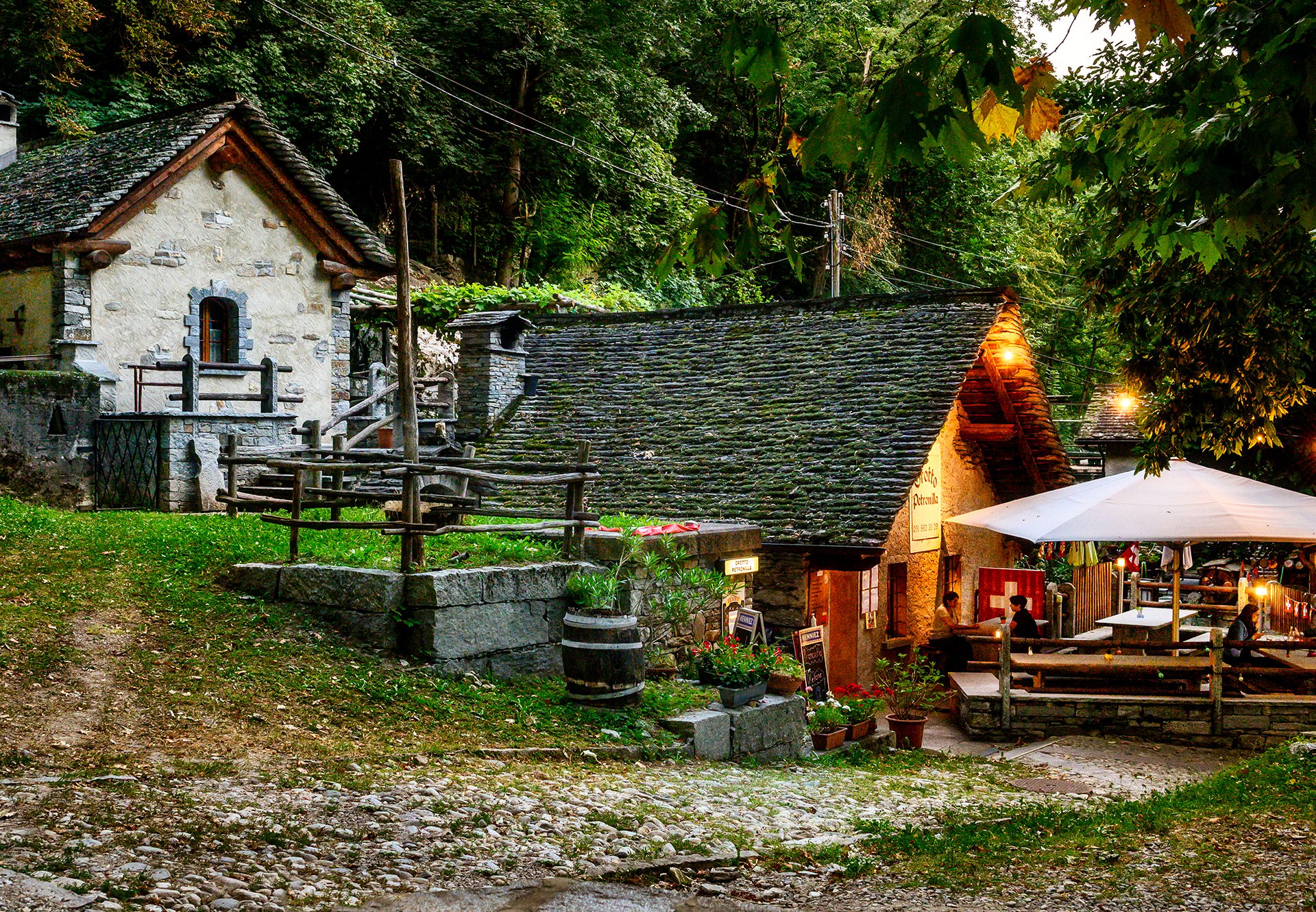 Atmospheric evening shot of the Grotto Petronilla in Biasca with traditional stone houses, illuminated terrace and cosy atmosphere surrounded by greenery.