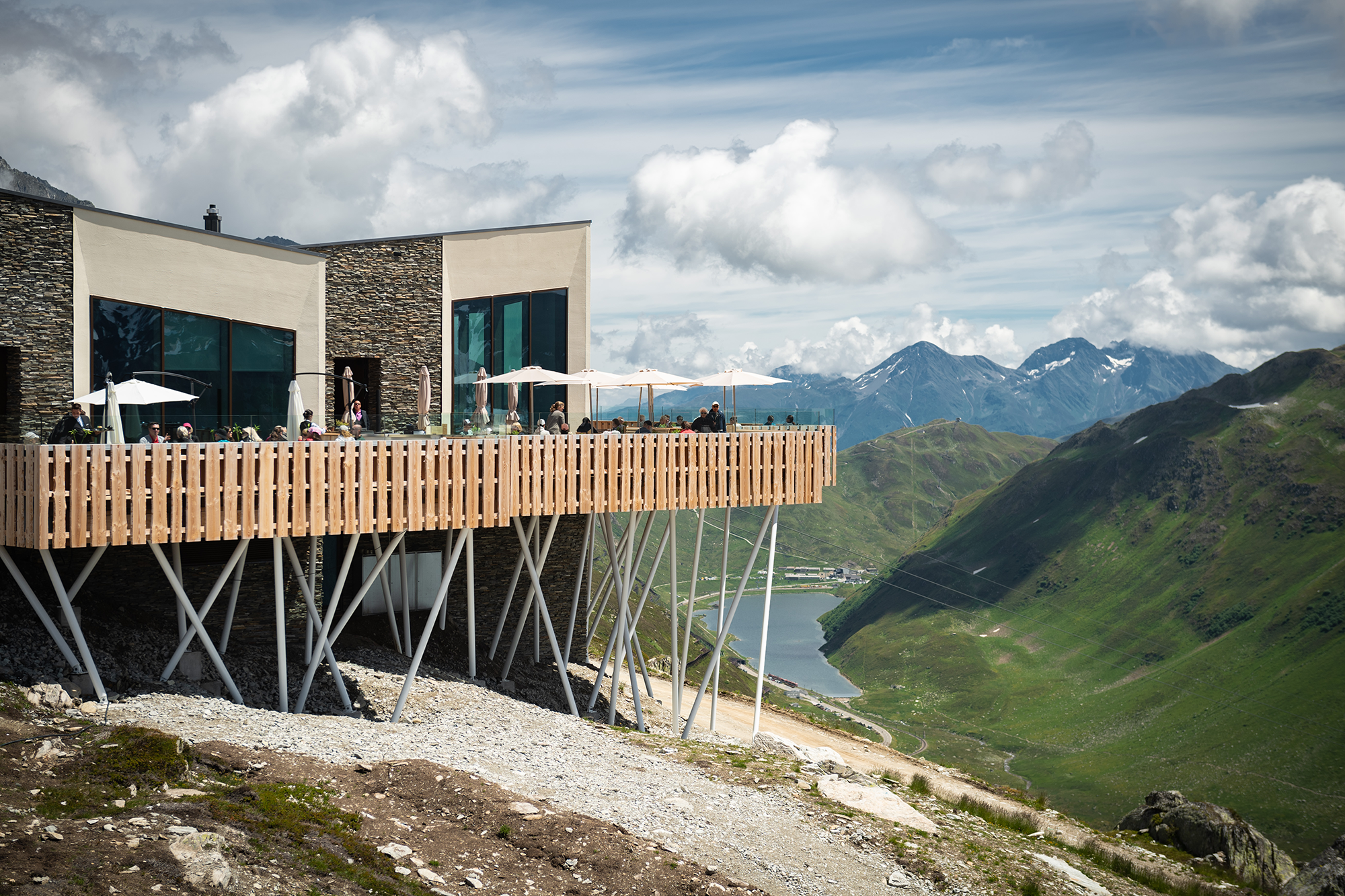 Das stilvolle Restaurant Gütsch mit grosser Sonnenterrasse thront auf 2300 m ü. M. über dem Tal – Gäste sitzen unter Sonnenschirmen, im Hintergrund das Bergpanorama und der Oberalpsee