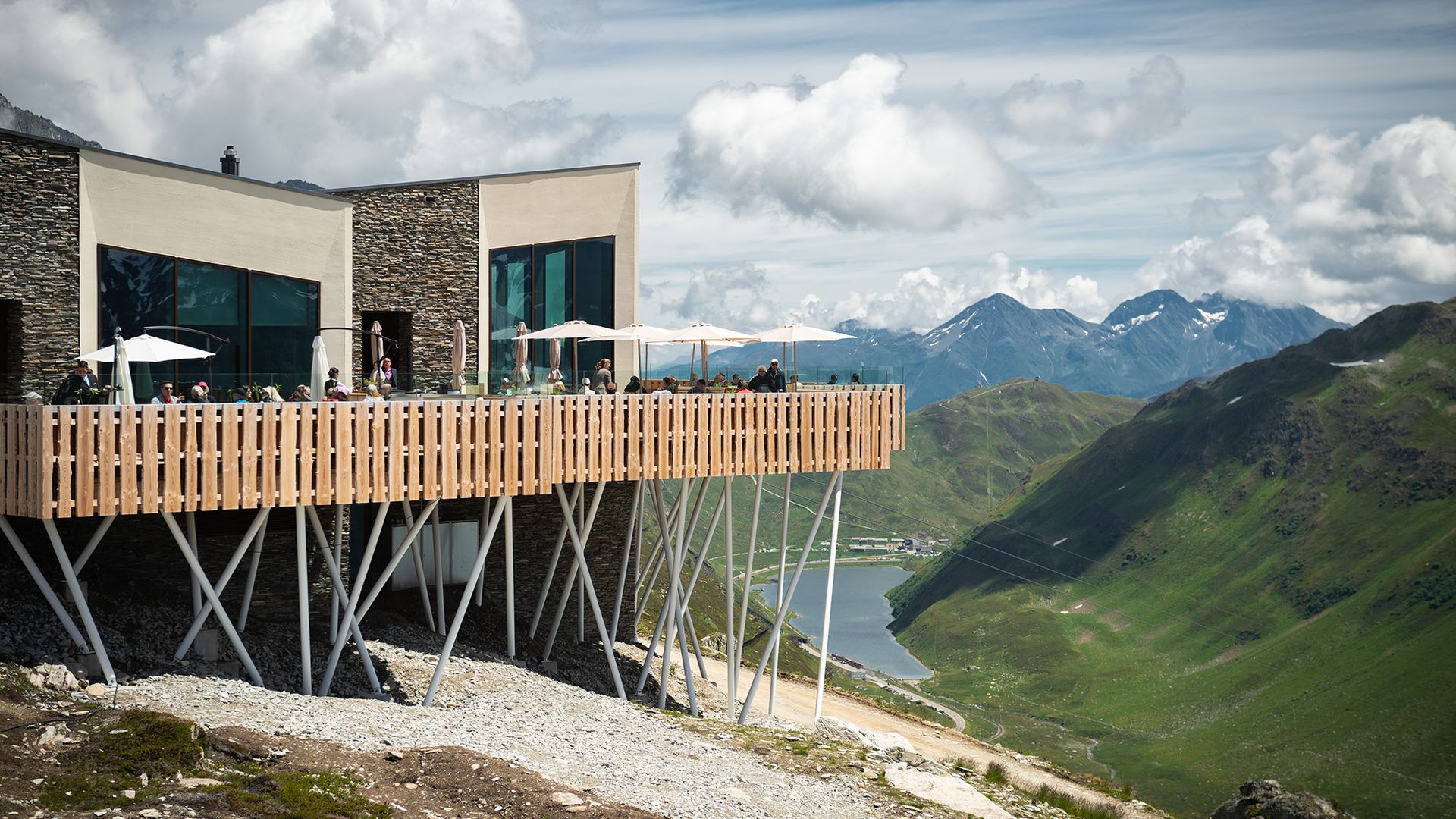 Il raffinato ristorante Gütsch con ampia terrazza soleggiata si trova a 2300 m di altitudine – ospiti seduti all’ombra degli ombrelloni, sullo sfondo le montagne e il lago di Oberalp