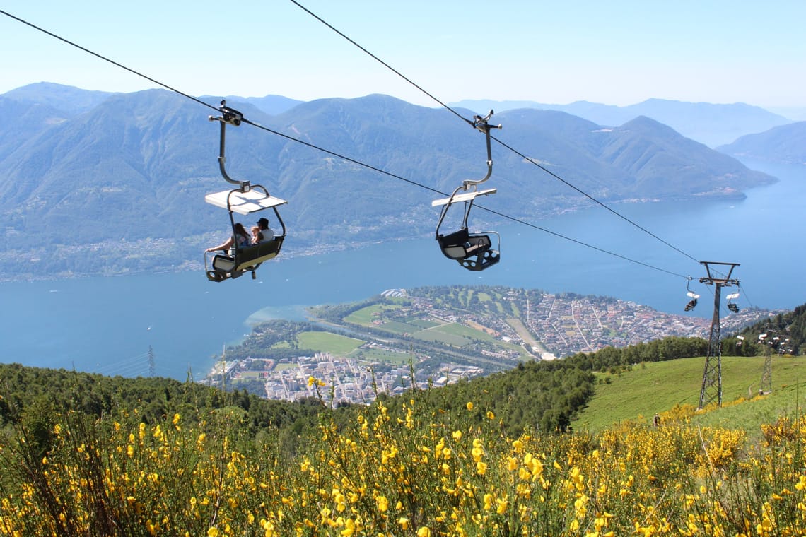 Zwei Sessellifte fahren entlang der Seilbahnstrecke über dem Lago Maggiore und an Blumenfeldern vorbei