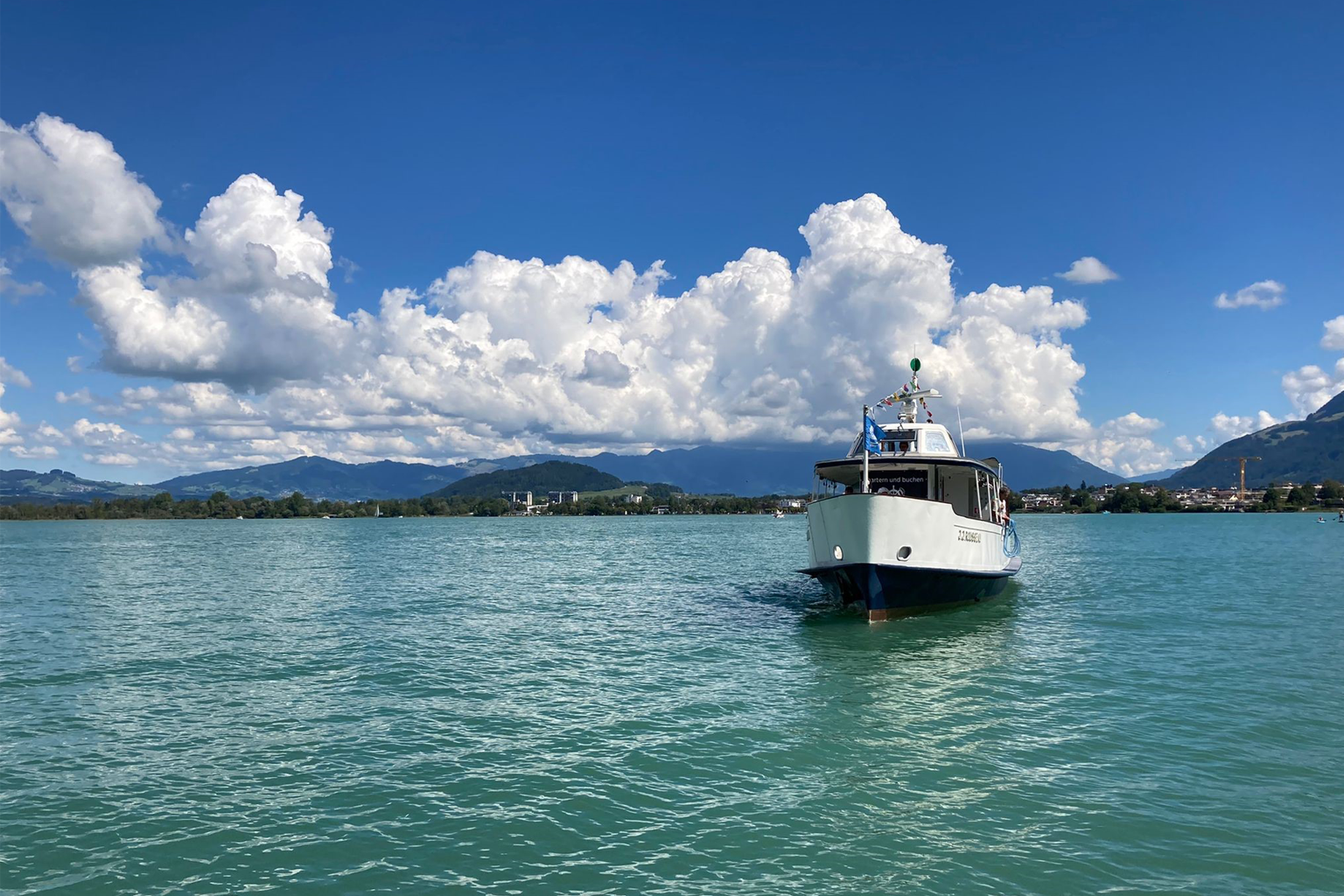 Il traghetto Obersee attraversa il Lago di Zurigo sotto un cielo estivo luminoso, con le colline circostanti e la città di Rapperswil sullo sfondo.
