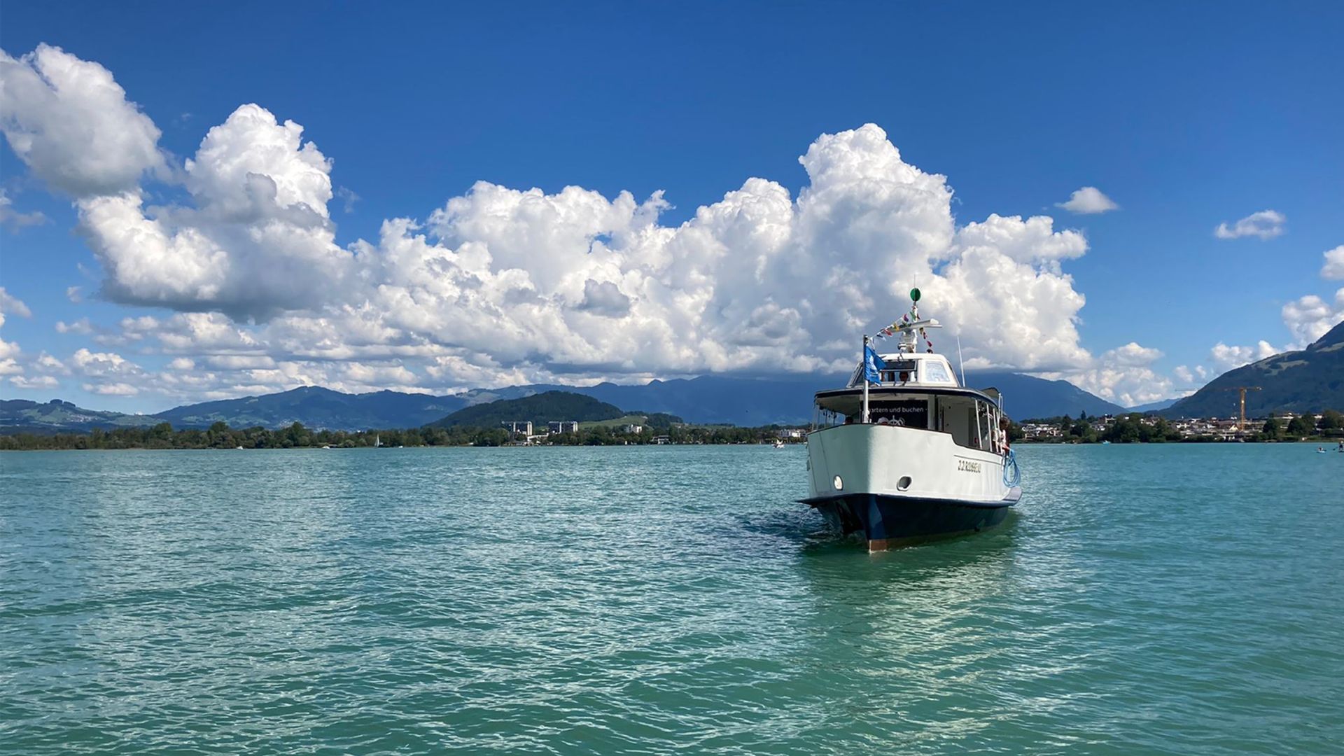 Die Oberseefähre fährt über den Zürichsee bei strahlendem Sommerwetter, mit Blick auf die umliegenden Hügel und die Stadt Rapperswil im Hintergrund.
