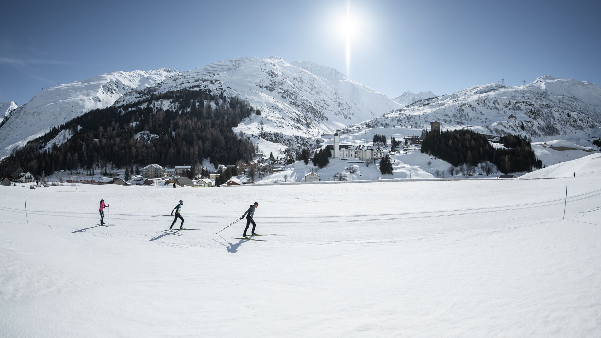 Trois skieurs de fond sur la piste à Andermatt avec vue sur l’église de Wassen et les montagnes enneigées.
