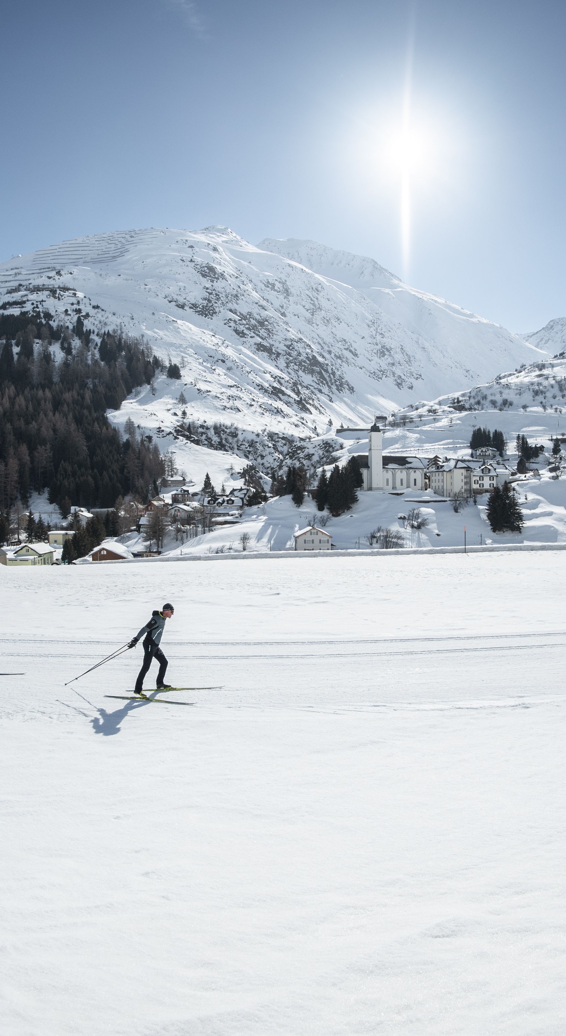 Three cross-country skiers on the trail in Andermatt with a view of the Wassen church and the snow-covered mountains.