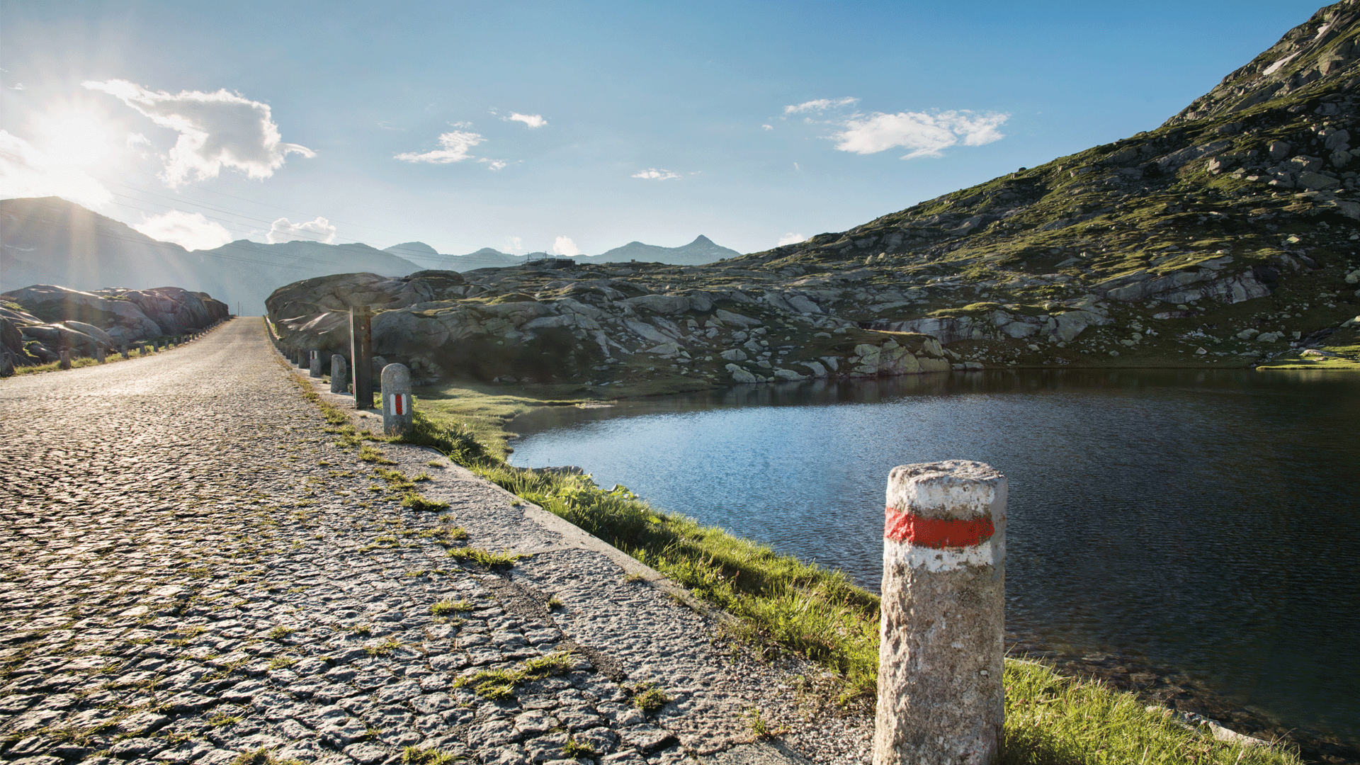 Sonnenlicht über der alten Kopfsteinpflasterstrasse am Gotthardpass mit Blick auf einen Bergsee und felsige Landschaft – Abschnitt der historischen Strada Alta oberhalb von Airolo im Tessin.