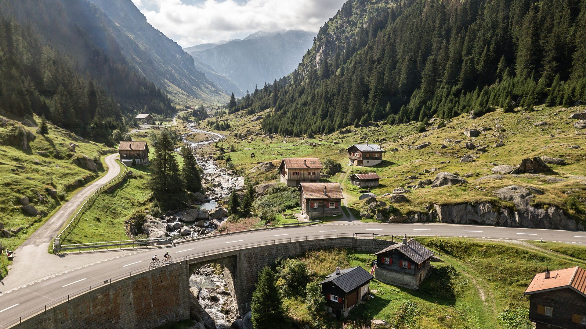 View of the Lukmanier Pass valley with a winding mountain road, wooden chalets, and cyclists surrounded by breathtaking Alpine scenery.