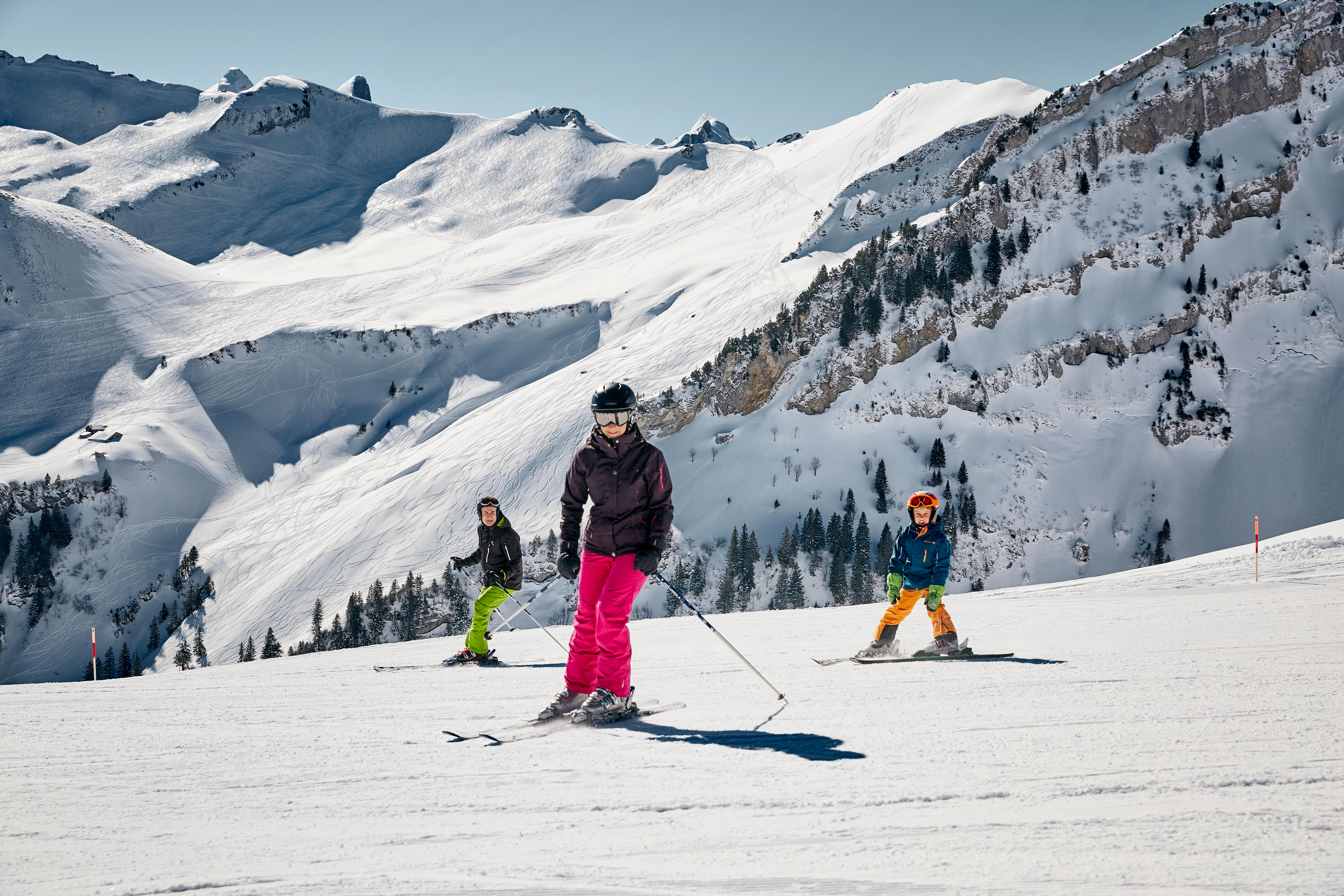 Drei Kinder mit farbigen Skihosen und dunkler Jacke fahren über eine Skipiste bei schönem Wetter