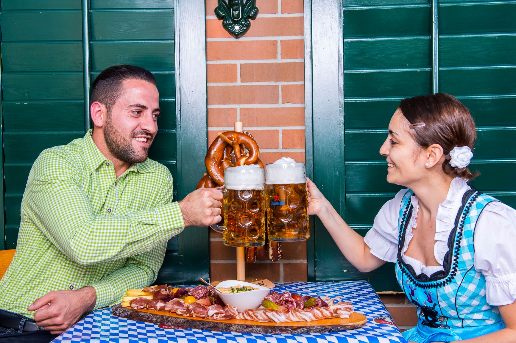 Mann und Frau stoßen beim Oktoberfest mit Maßkrügen Bier an, vor ihnen eine Brotzeitplatte mit Wurst, Käse und Brezeln.