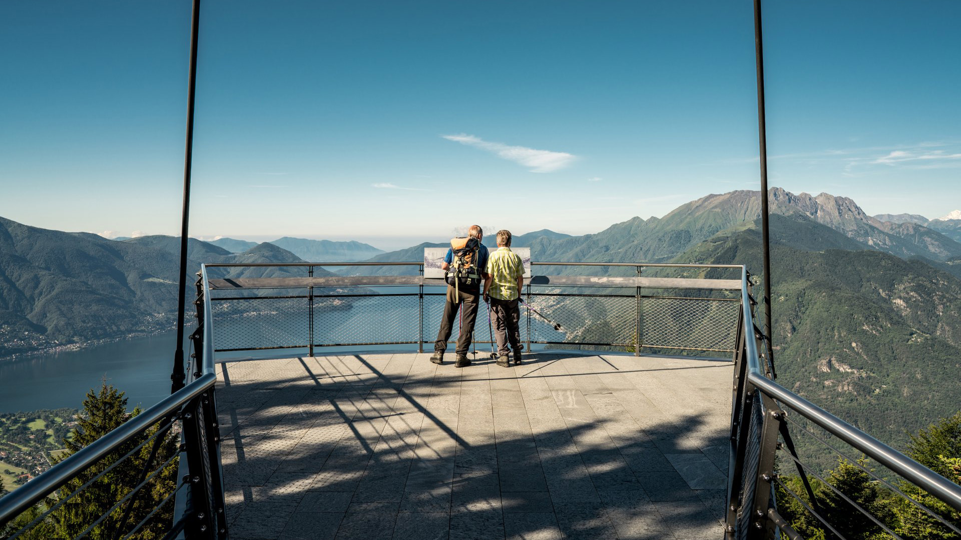 Due escursionisti sulla piattaforma panoramica di Cardada Cimetta con vista sul Lago Maggiore e sulle montagne del Ticino sotto un cielo sereno.