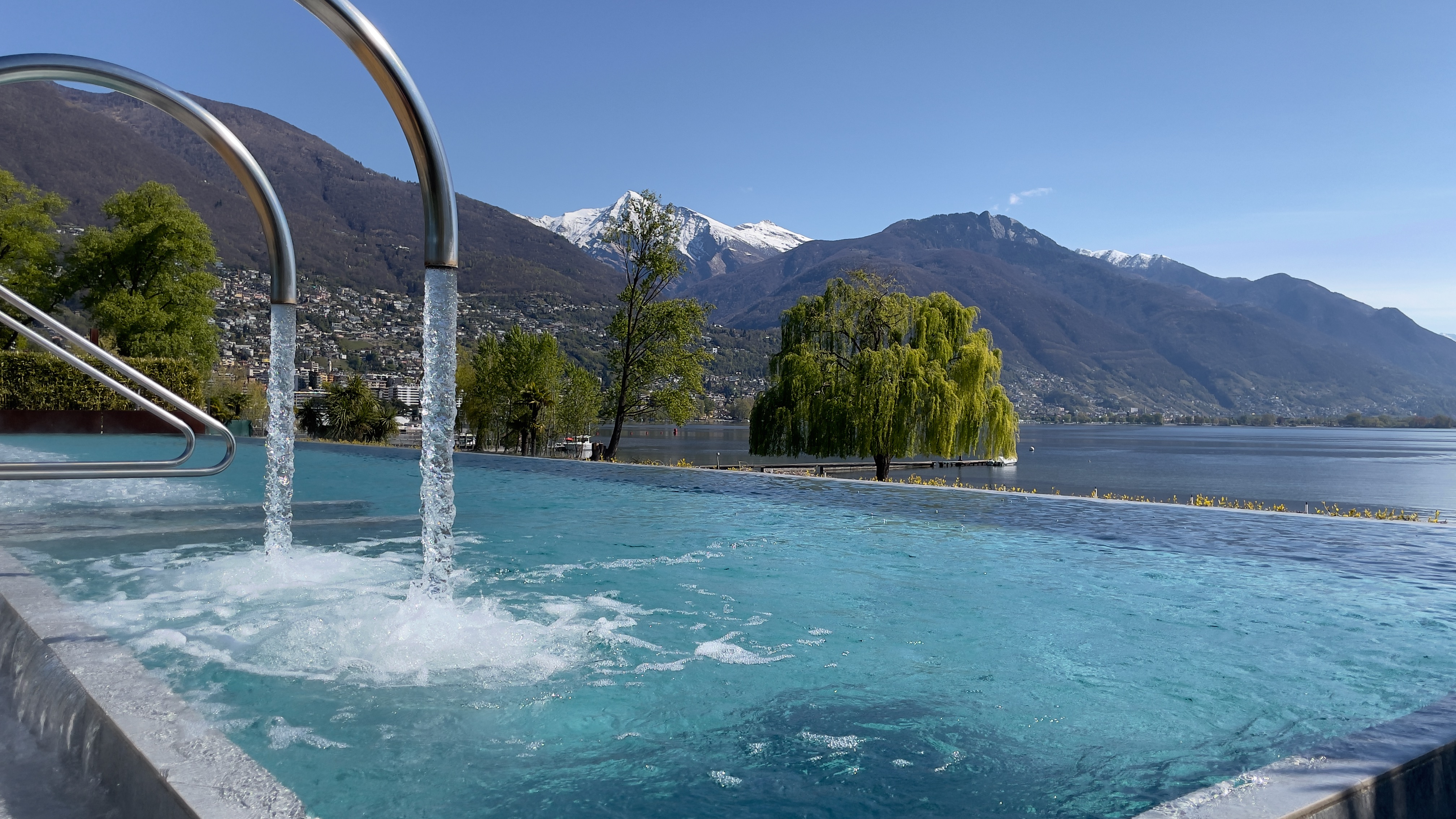Aussenbecken der Termali Salini & Spa Locarno mit sprudelndem Wasser und Panoramablick auf den Lago Maggiore, umgeben von Bergen und grünen Weiden.