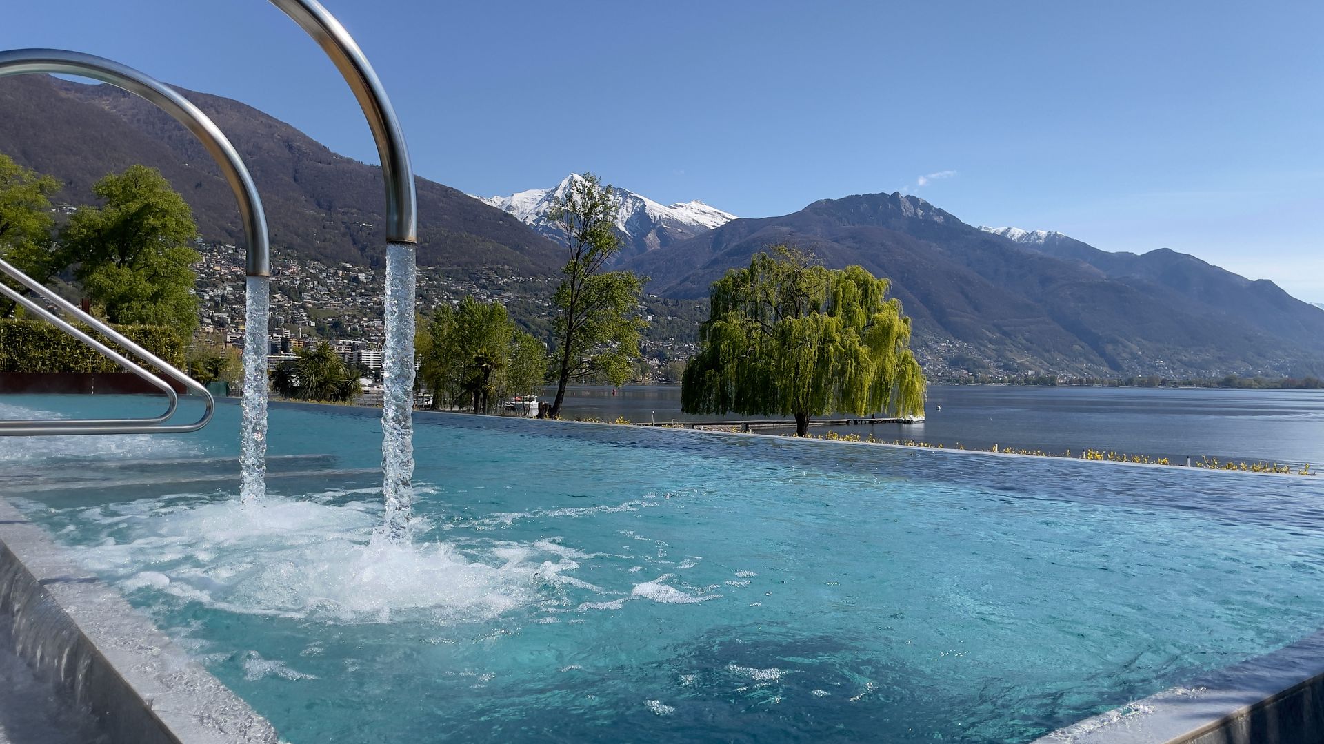Outdoor pool of Termali Salini & Spa Locarno with bubbling water and panoramic view of Lake Maggiore, surrounded by mountains and lush greenery.