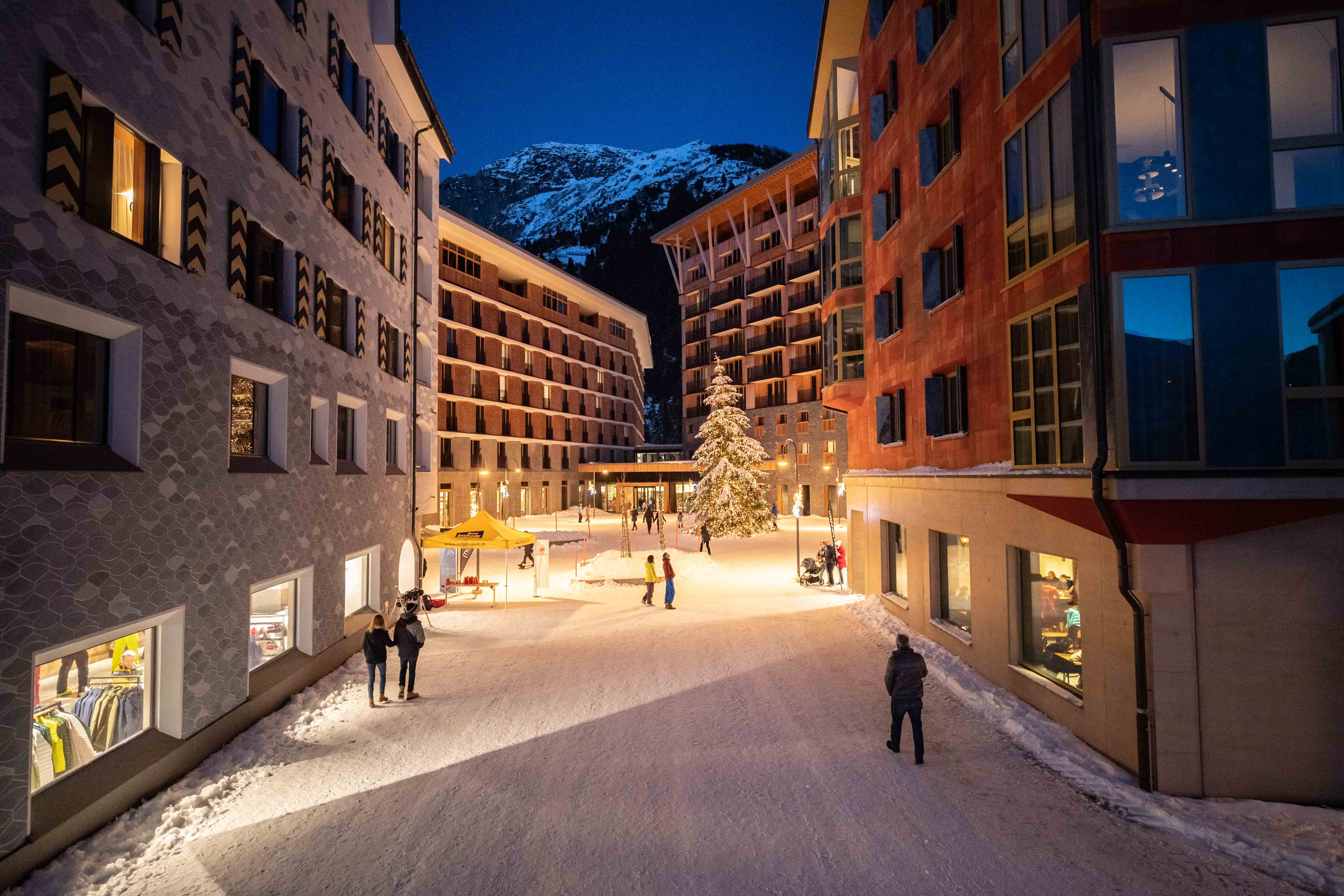 Winterliche Piazza Gottardo in Andermatt mit beleuchtetem Weihnachtsbaum, umgeben von mehrstöckigen Gebäuden und schneebedecktem Boden bei Abenddämmerung.
