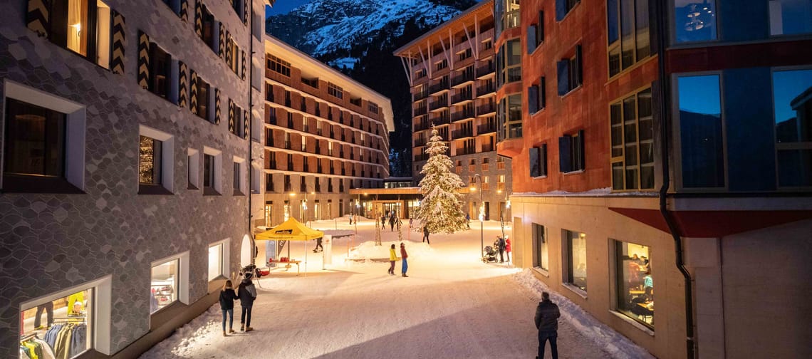 Winterliche Piazza Gottardo in Andermatt mit beleuchtetem Weihnachtsbaum, umgeben von mehrstöckigen Gebäuden und schneebedecktem Boden bei Abenddämmerung.