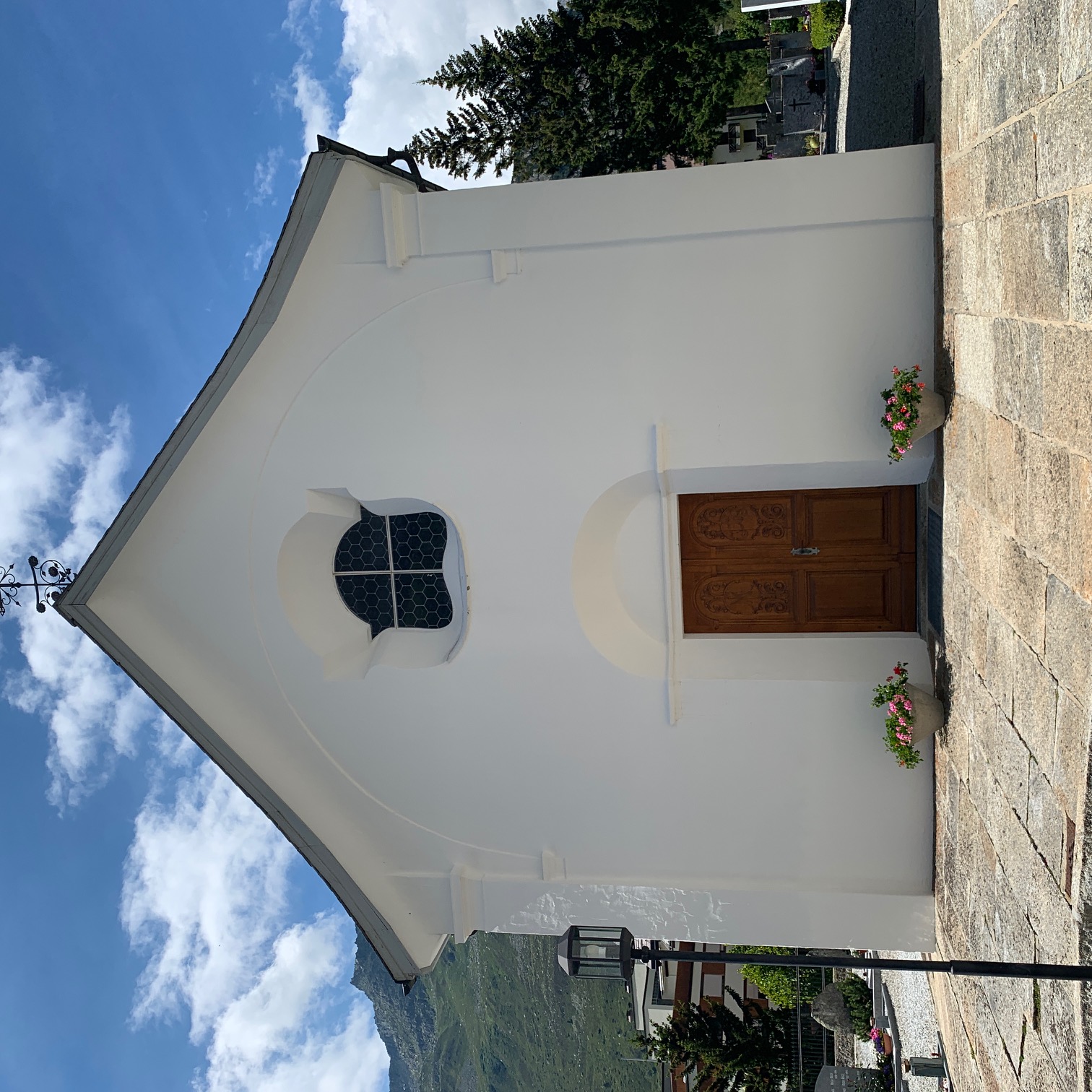 Weisse Kapelle St. Michael in Andermatt mit brauner Holztür, kleinem Fenster mit Rundbogen und zwei Blumentöpfen davor.