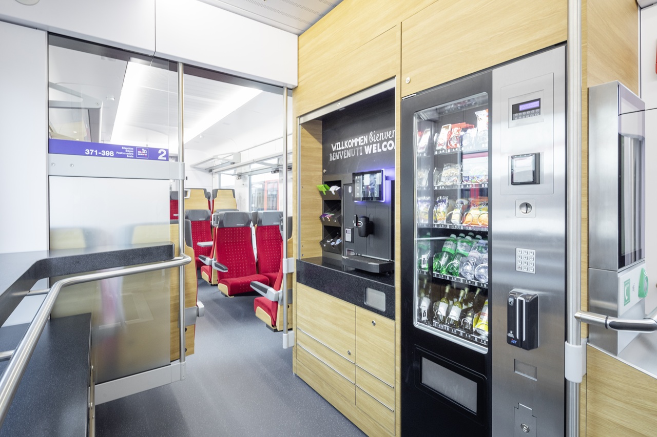 Innenansicht eines modernen Zugwagens mit Snack- und Getränkeautomaten sowie Kaffeemaschine; daneben Glaswand und Durchgang zu Sitzplätzen mit roten Sitzen, darüber ein „Willkommen / Bienvenue / Welcome“-Hinweis.