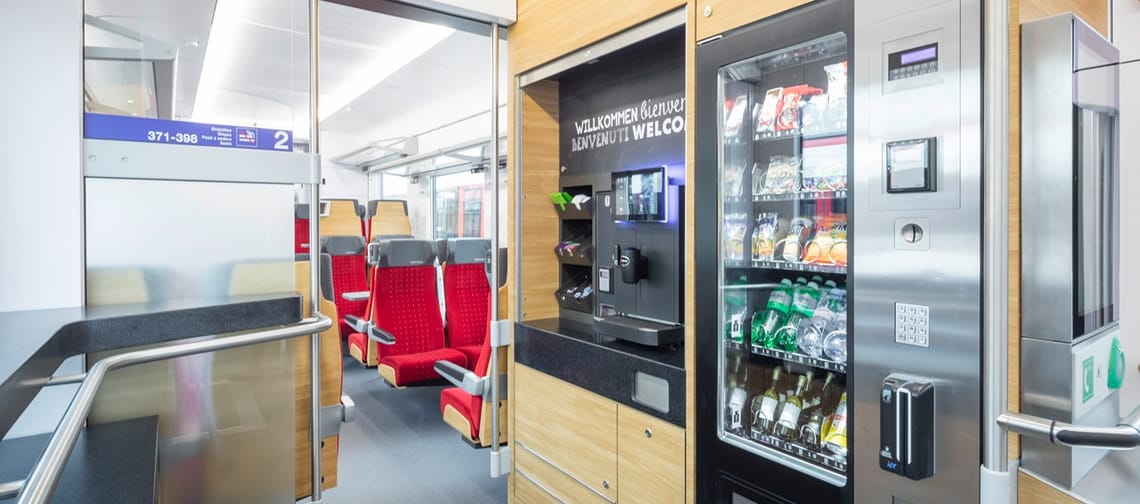 Innenansicht eines modernen Zugwagens mit Snack- und Getränkeautomaten sowie Kaffeemaschine; daneben Glaswand und Durchgang zu Sitzplätzen mit roten Sitzen, darüber ein „Willkommen / Bienvenue / Welcome“-Hinweis.