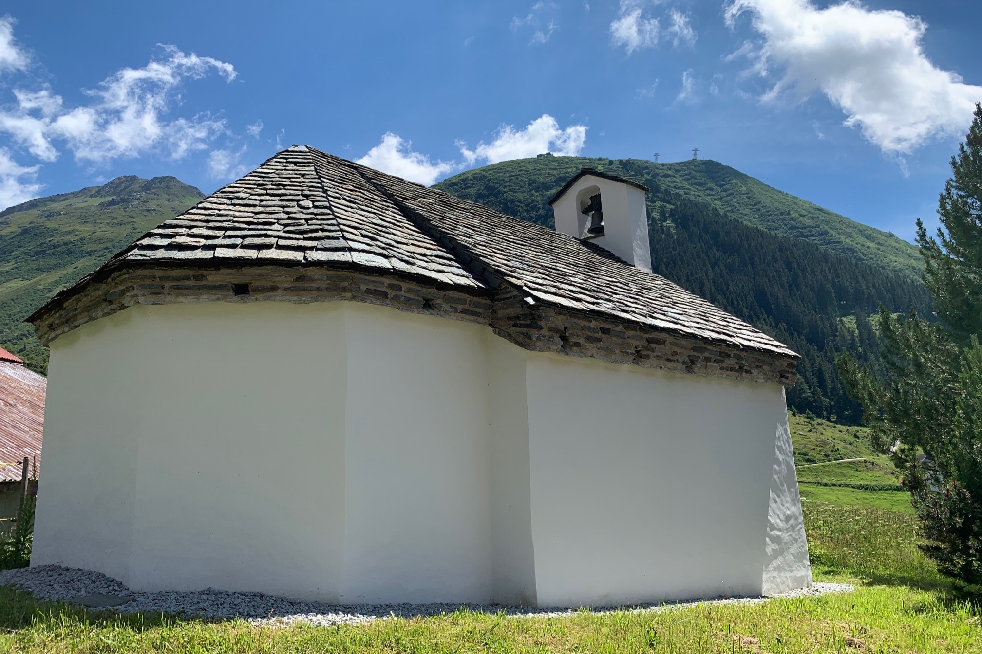 Chapel of St. Wendelin viewed from the rear with white plastered walls, stone shingle roof, and small bell tower against a forested mountain backdrop