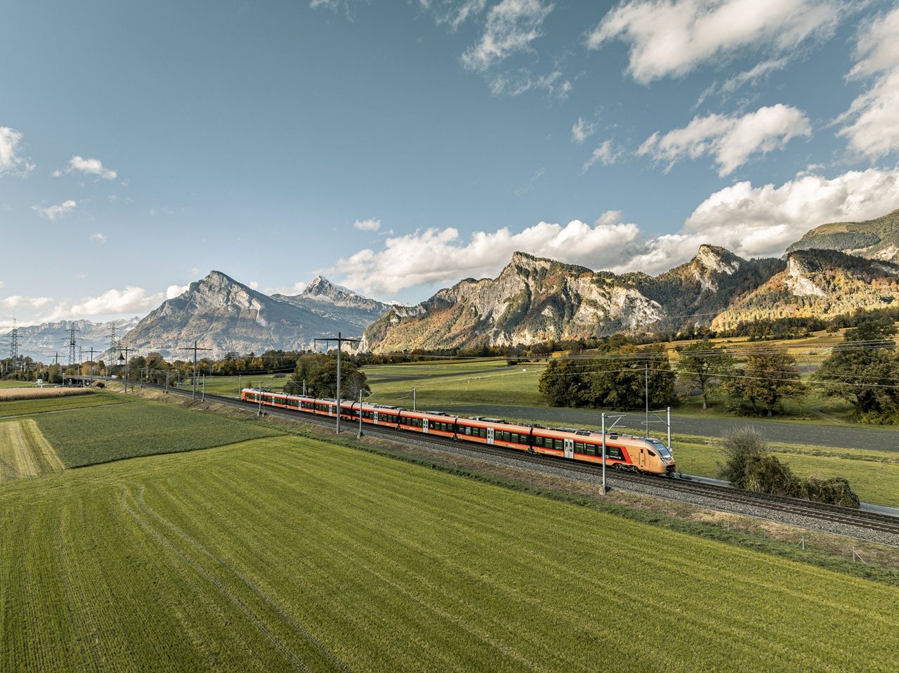 Ein Zug fährt auf Gleisen durch eine grüne Wiese vor einer Berglandschaft. Darüber erstreckt sich ein blauer Himmel mit weissen Wolken.