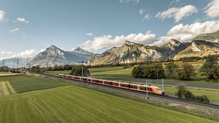 Ein Zug fährt auf Gleisen durch eine grüne Wiese vor einer Berglandschaft. Darüber erstreckt sich ein blauer Himmel mit weissen Wolken.