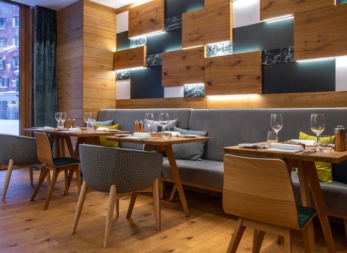 Restaurantbereich mit Holztischen, gepolsterten Stühlen und einer Wand mit beleuchteten Holz- und Steinpaneelen im Radisson Blu Hotel Reussen in Andermatt