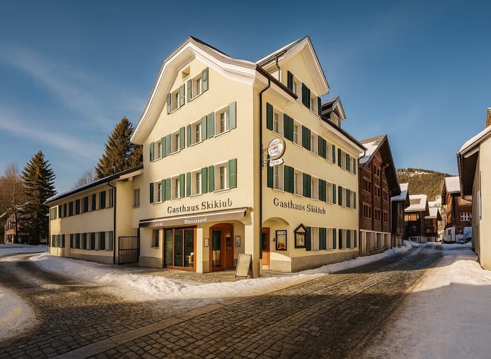 Dreigeschossiges Gasthaus mit grün lackierten Fensterläden und Schriftzug »Gasthaus Skiklub« an der Fassade in verschneiter Dorfstrasse