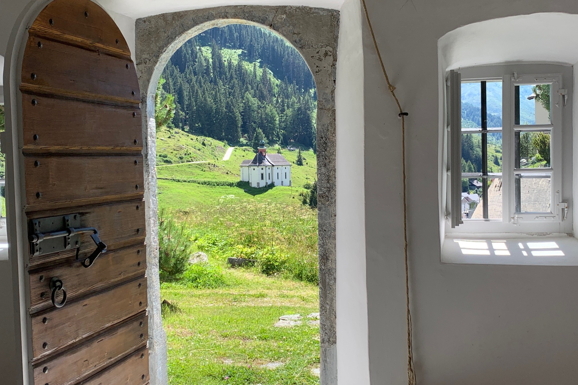 Vue depuis l'intérieur d'une chapelle à travers une porte arrondie en pierre ouverte sur une chapelle blanche dans un paysage montagneux verdoyant avec forêt en arrière-plan