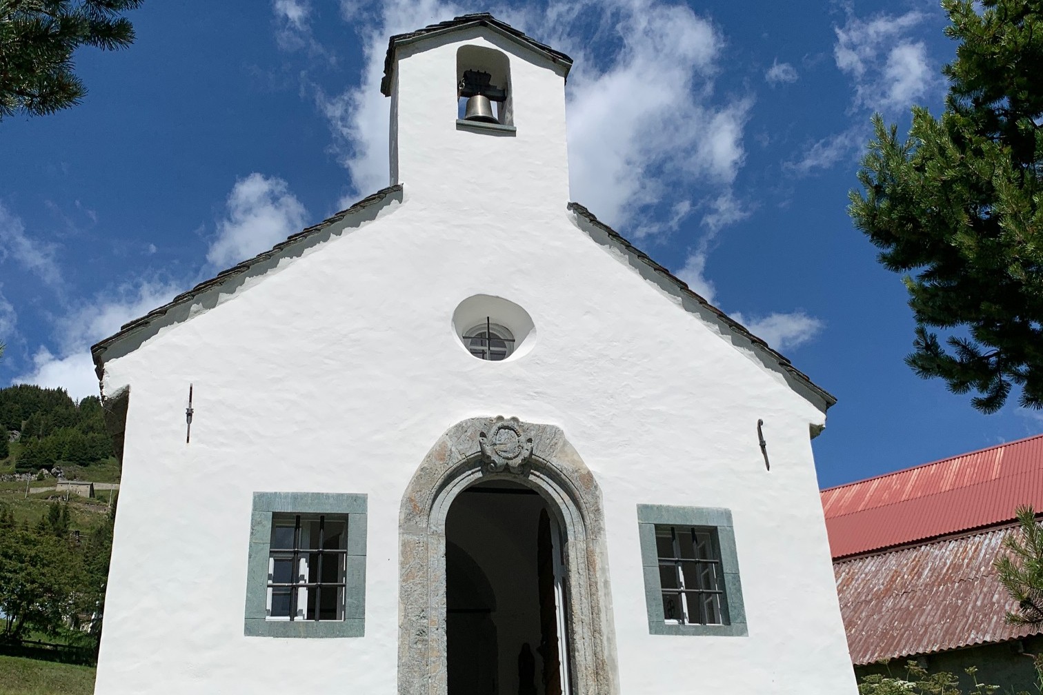Chapelle blanche Saint-Wendelin à Andermatt avec petit clocher deux fenêtres et entrée ouverte sous ciel bleu