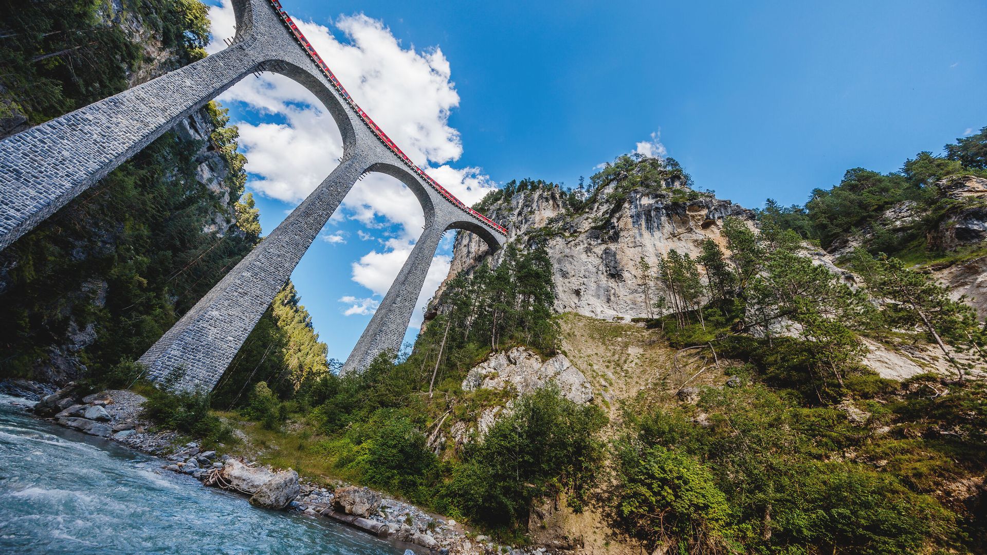 Über den Landwasserviadukt fährt ein roter RhB Zug und darunter fliesst der Fluss.