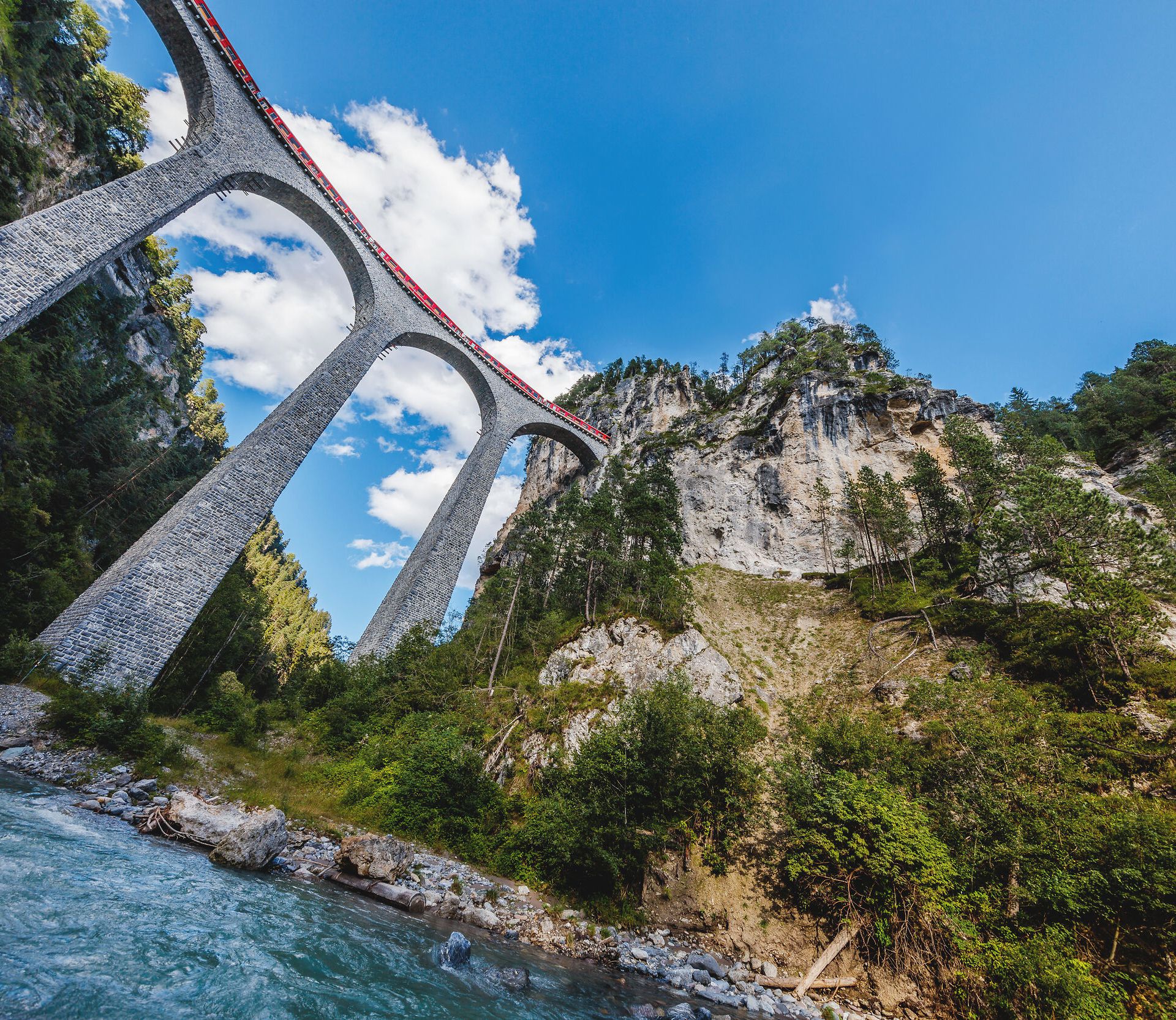 Über den Landwasserviadukt fährt ein roter RhB Zug und darunter fliesst der Fluss.