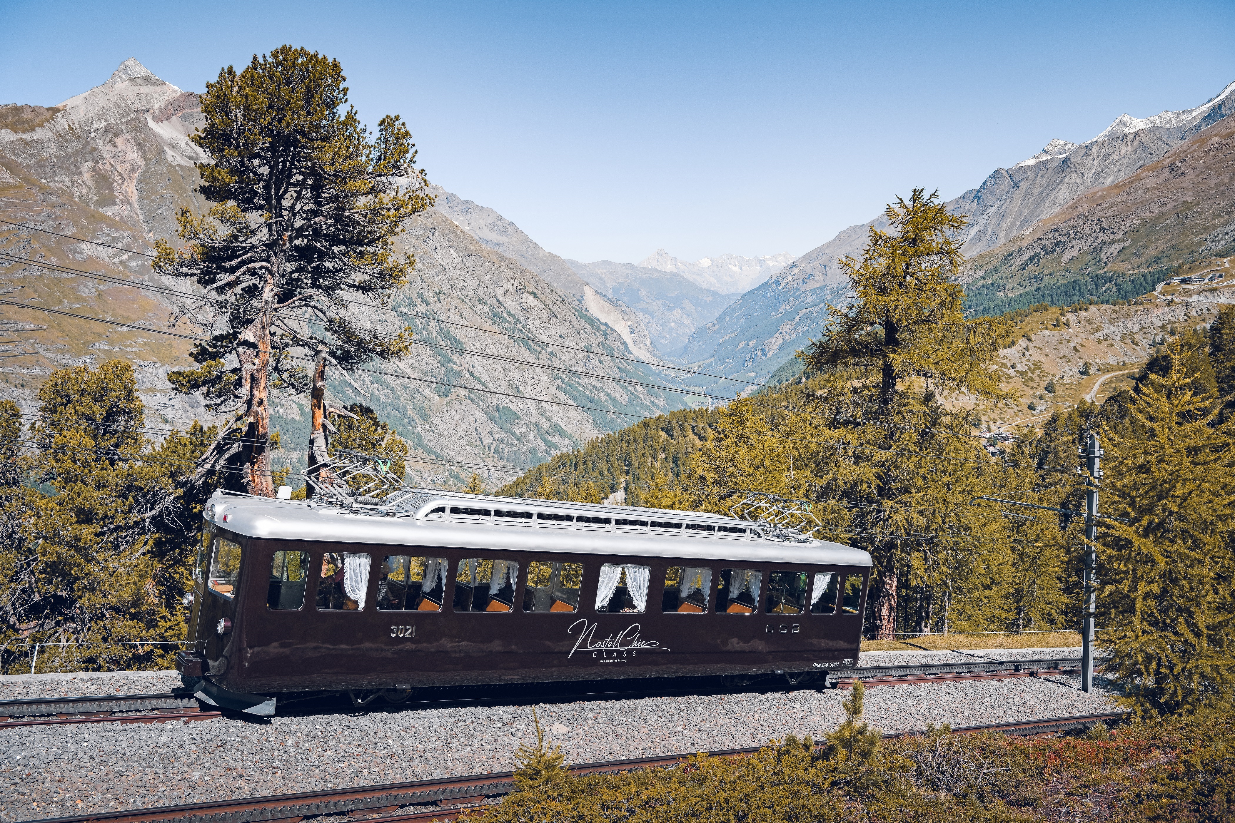  Le wagon NostalChic Class, entouré de forêt, roule sous un ciel bleu éclatant lors d'une journée d'été.