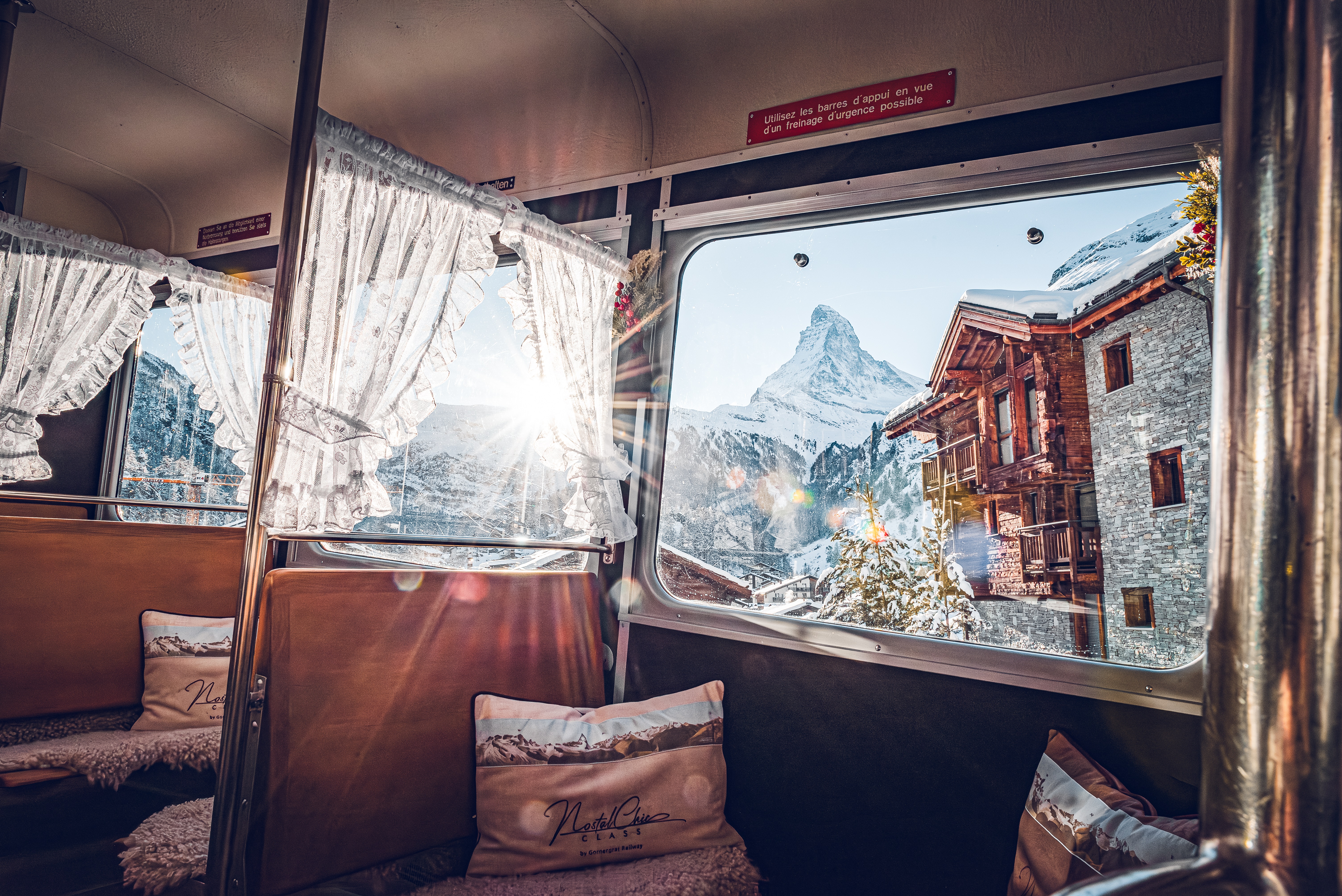 The NostalChic Class carriage, decorated with cushions and curtains, offers a warm ambience. Through the glass, the Matterhorn is revealed under a blue winter sky.

