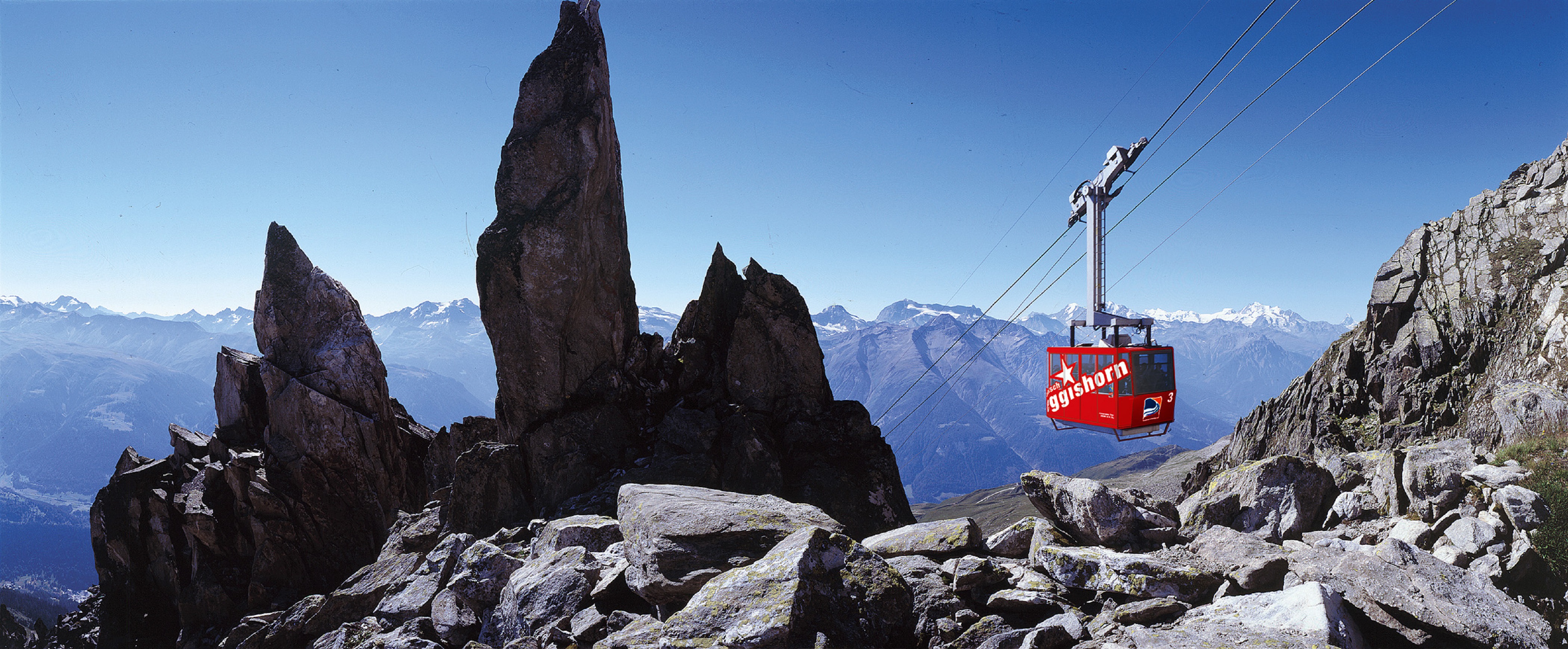  The red Eggishorn aerial tramway soars through a mountainous landscape under a clear, cloudless blue sky.
