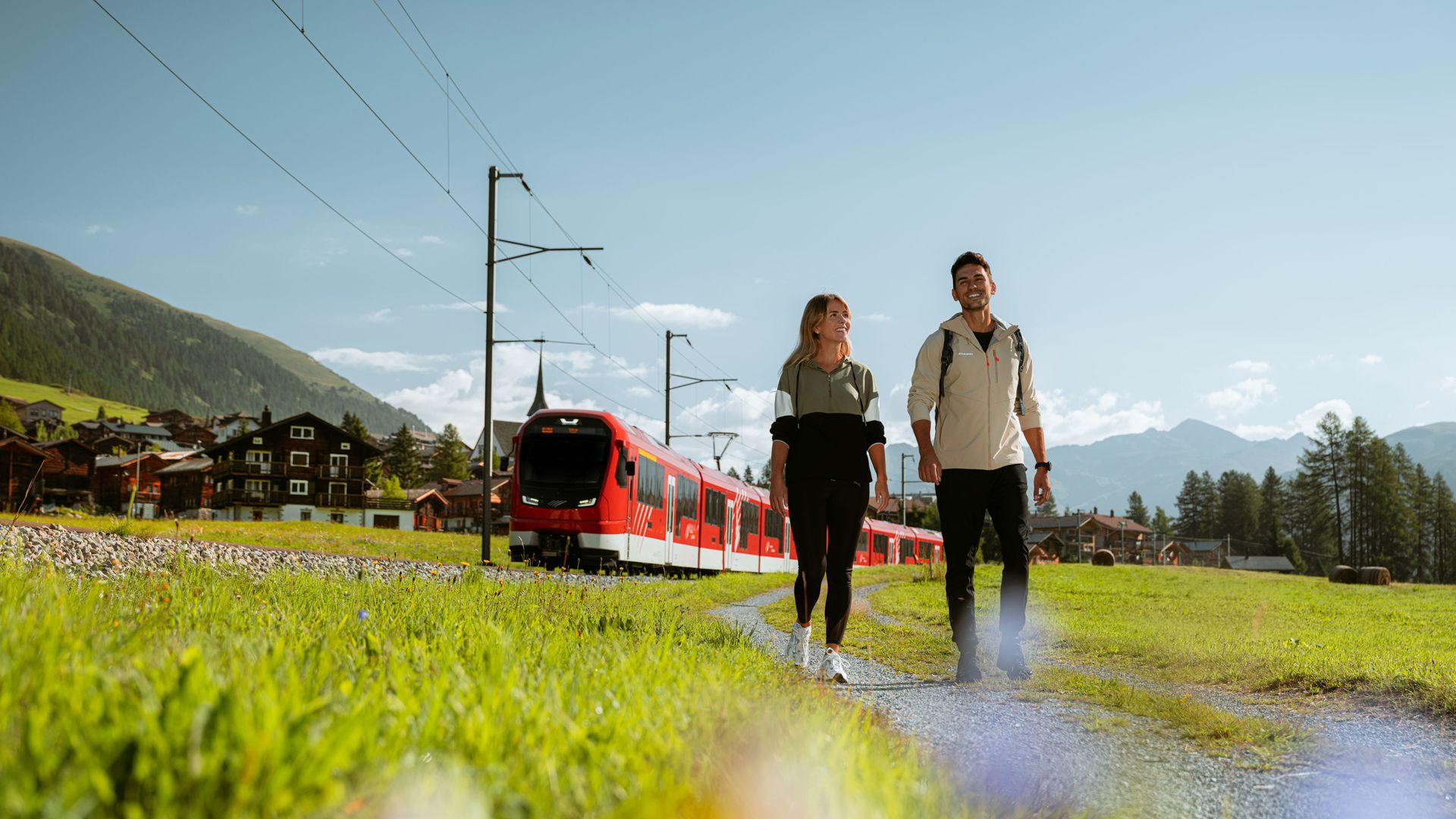 A couple stroll along a path in Goms, next to the Matterhorn Gotthard Bahn ORION train, on a sunny summer's day.