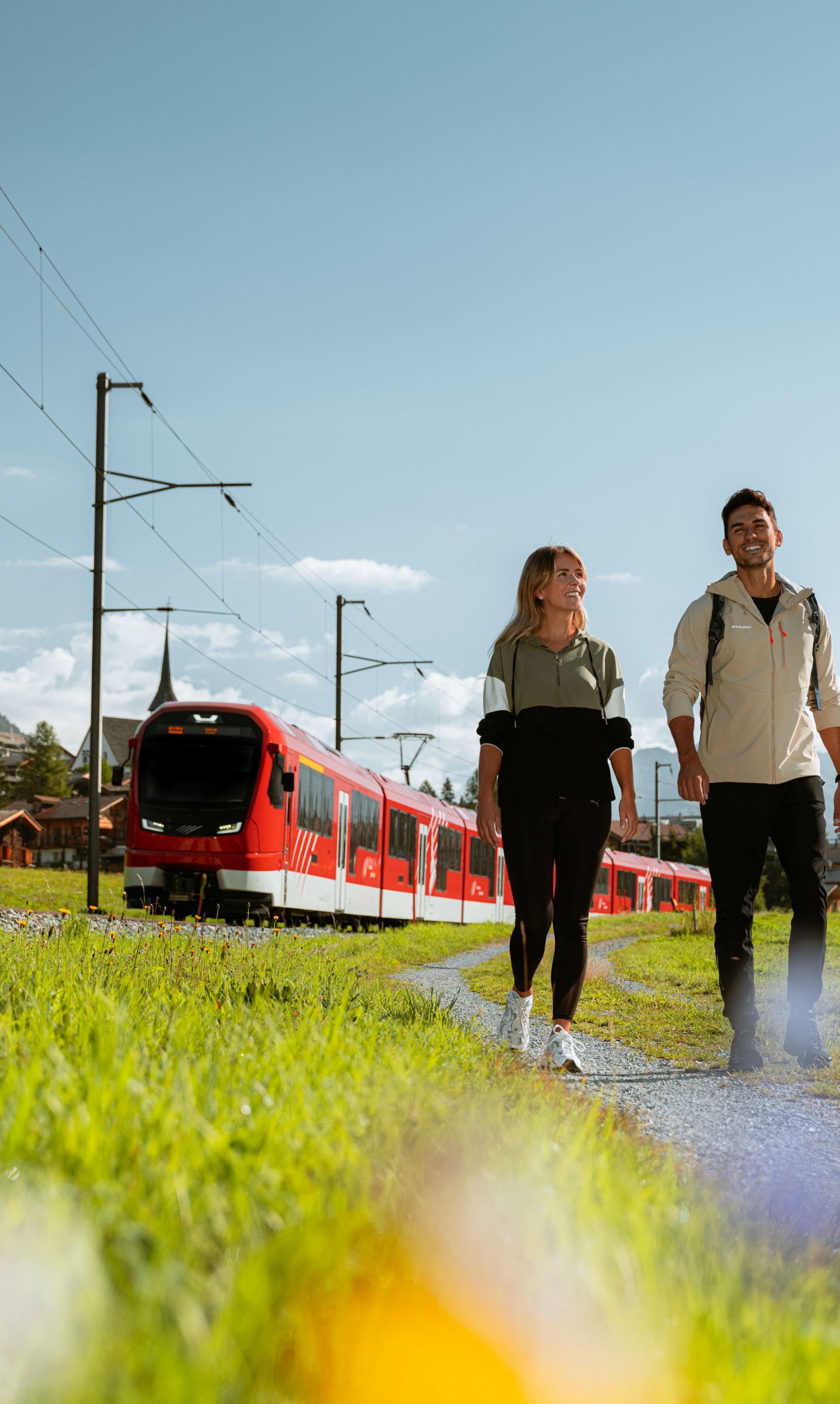 Un couple se promène sur un chemin dans la vallée de Conches, à côté du train ORION du Matterhorn Gotthard Bahn, lors d'une journée estivale ensoleillée.