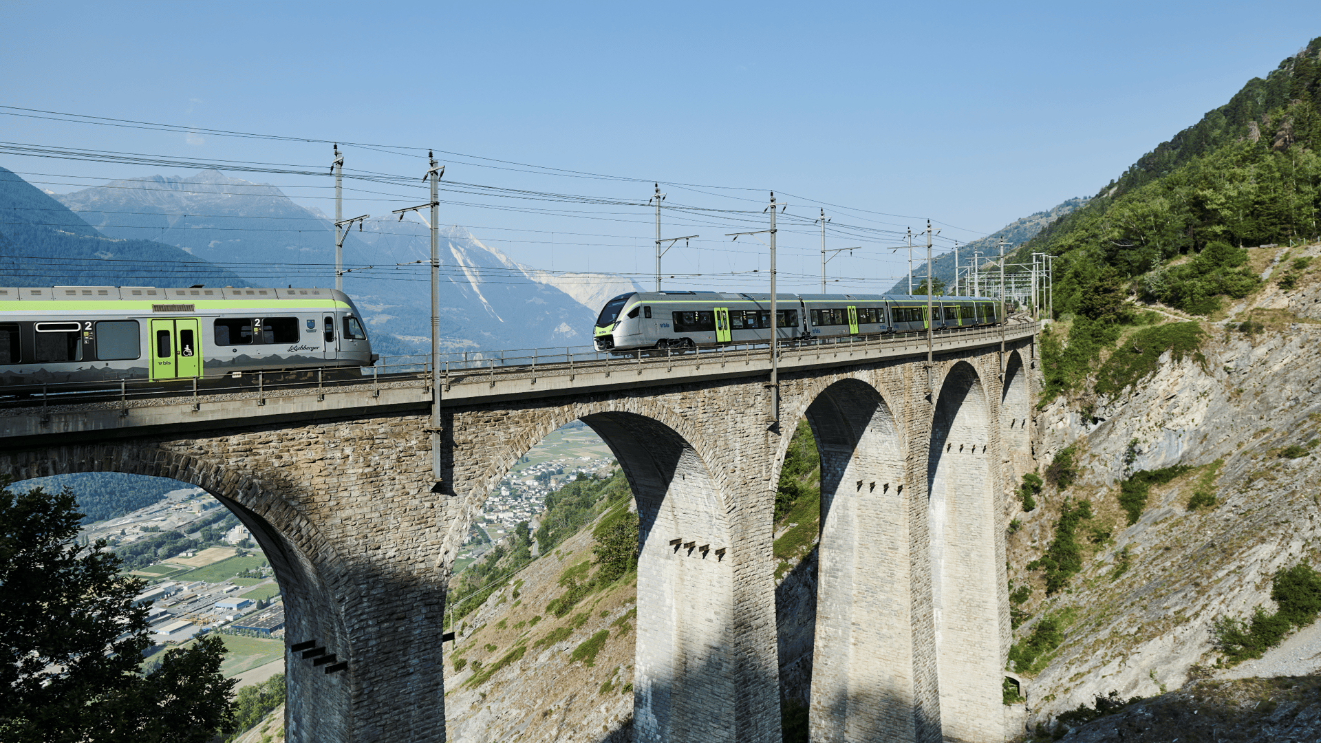 BLS-Züge fahren an einem sonnigen Sommertag unter einem wolkenlosen blauen Himmel über eine Brücke.
