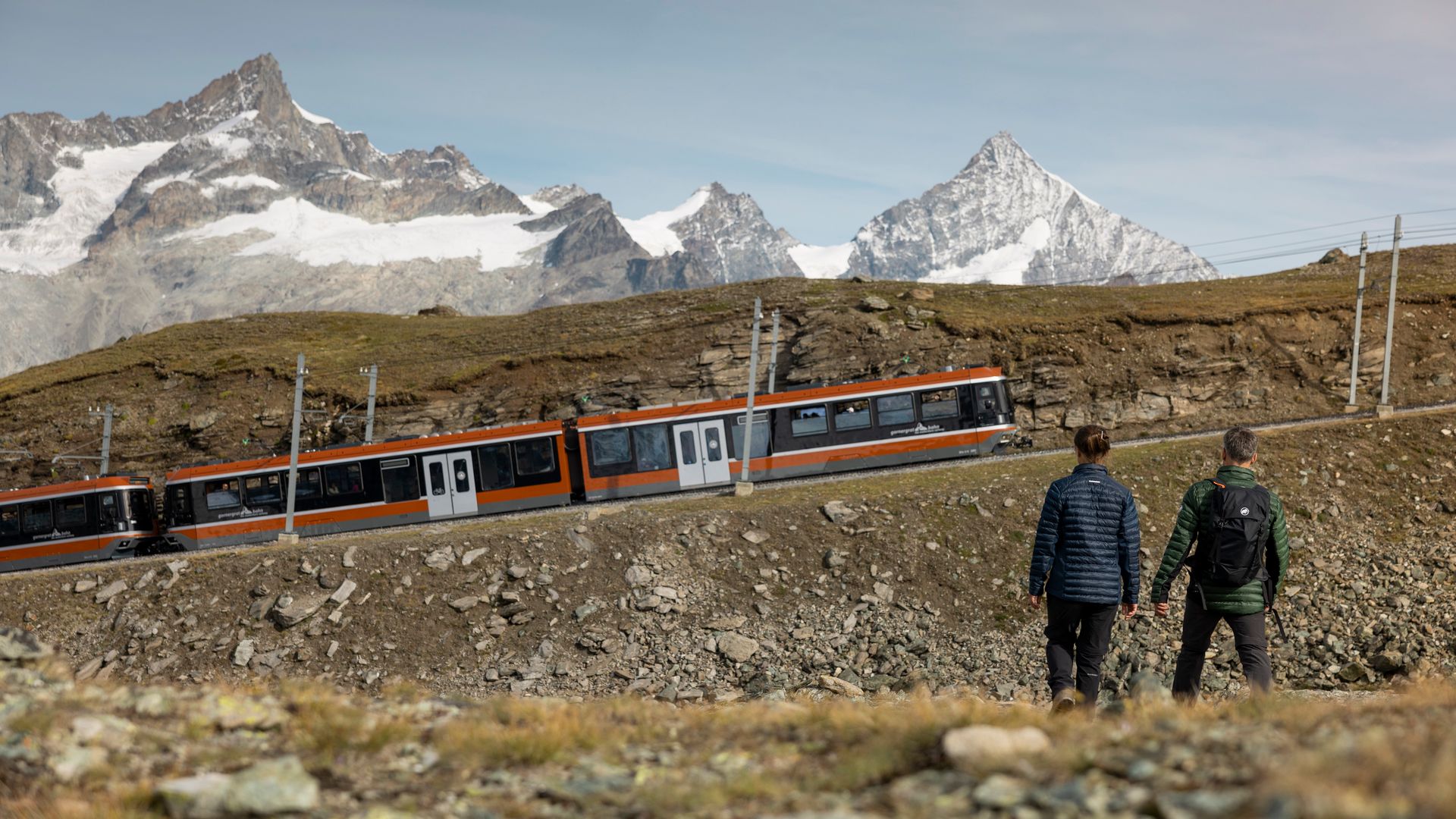 Deux hommes font de la randonnée à Zermatt tandis que le Gornergrat Bahn passe devant eux, lors d'une journée ensoleillée.