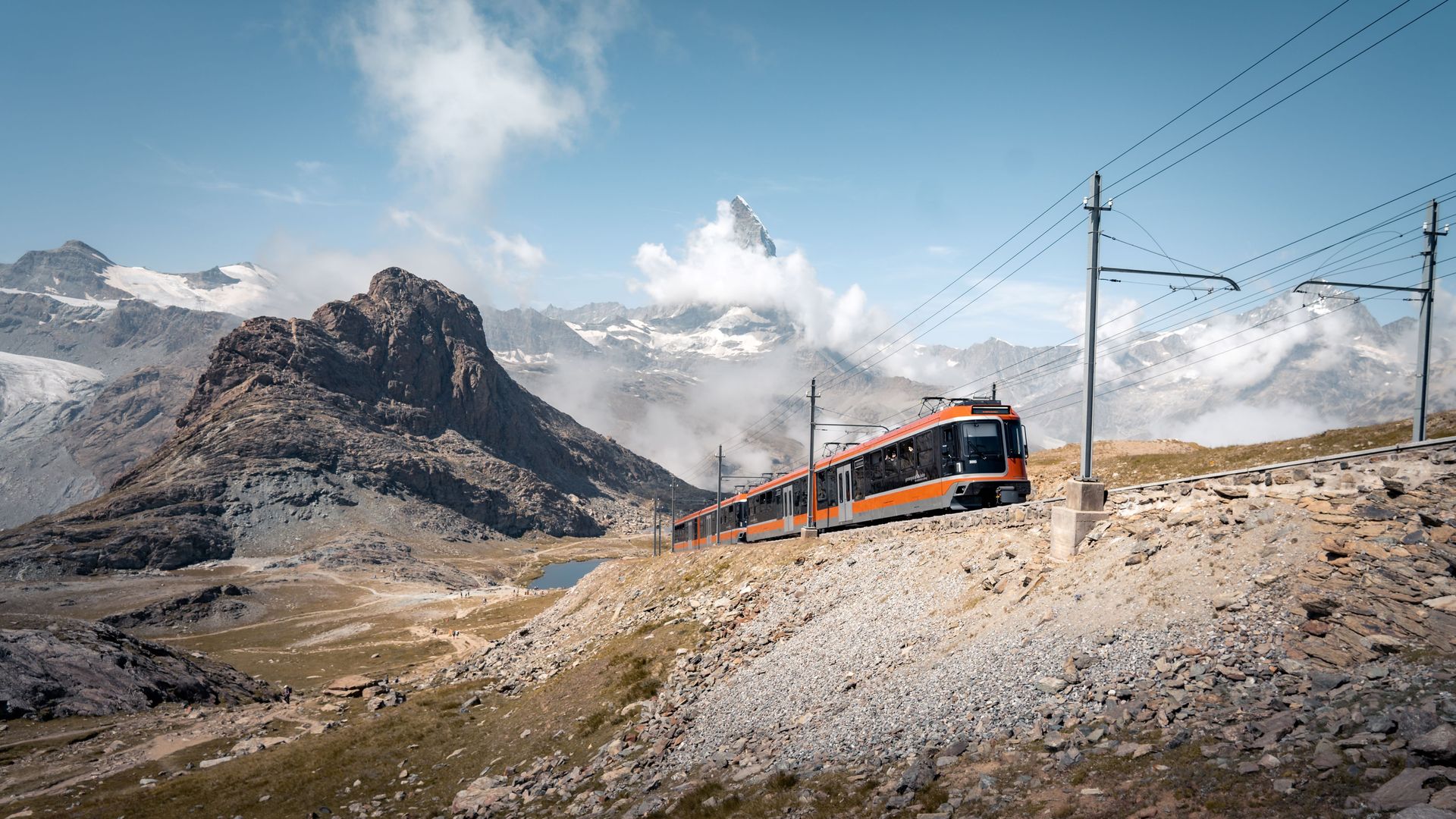 The Polaris train crosses a mountainous landscape on a summer's day, with the Matterhorn in the background, partially concealed by clouds and the Riffelsee.