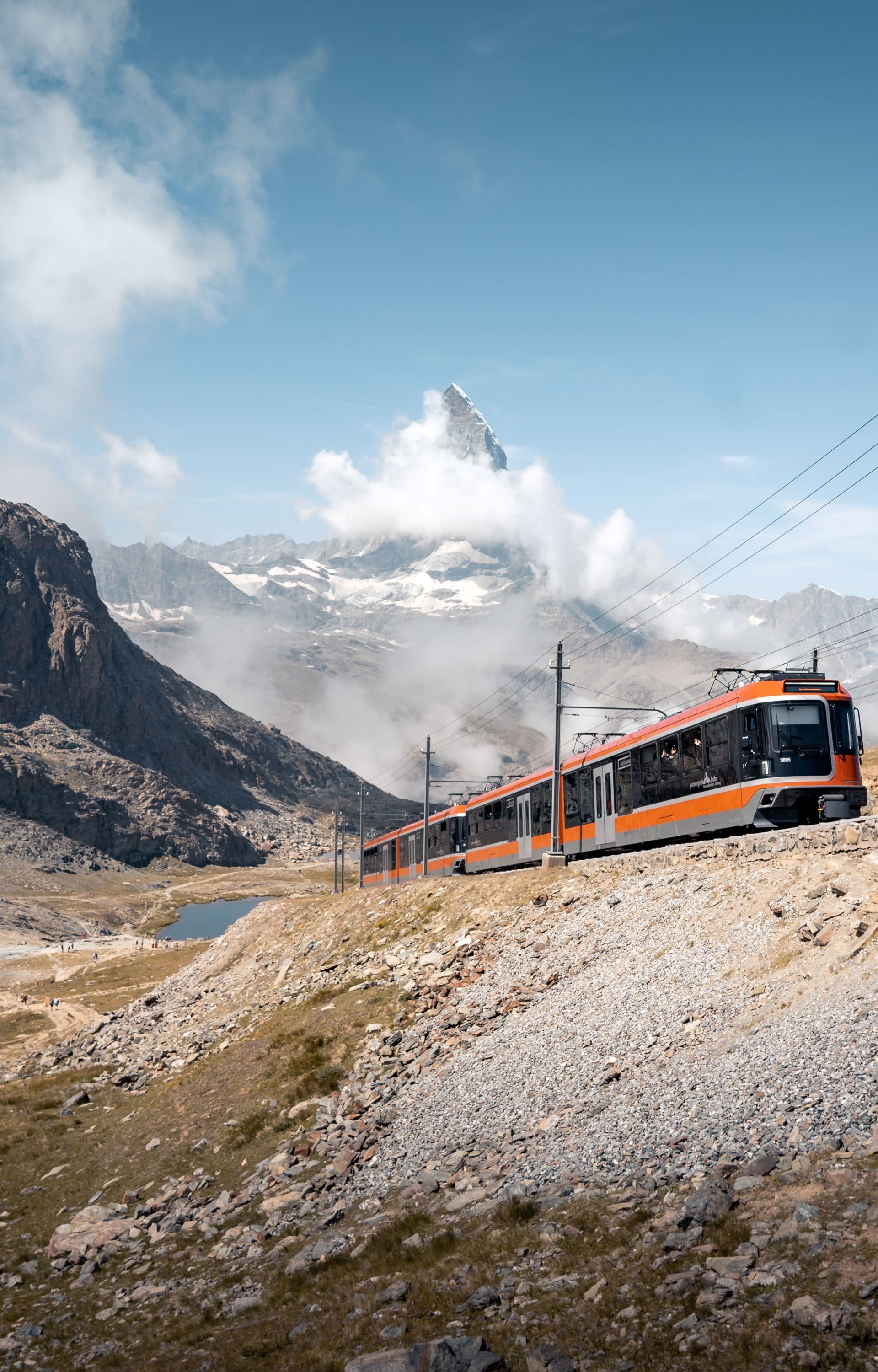 The Polaris train crosses a mountainous landscape on a summer's day, with the Matterhorn in the background, partially concealed by clouds and the Riffelsee.