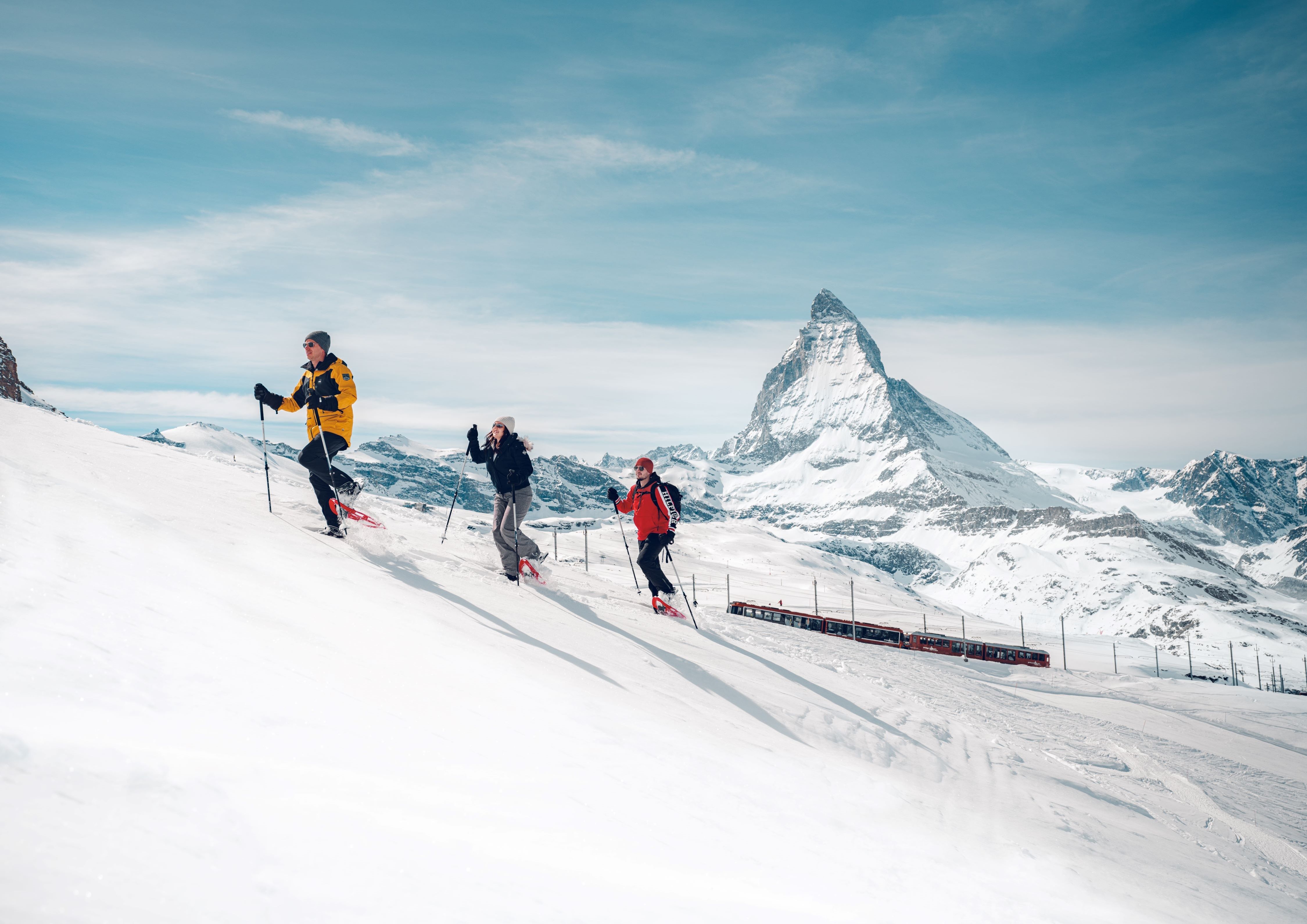 Three people walk on snowshoes through a snow-covered landscape, with the Gornergrat Bahn and the Matterhorn in the background.