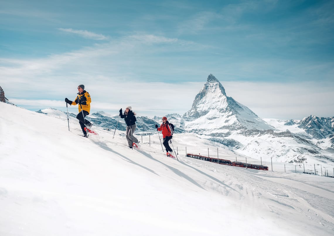 Schneeschuhwandern am Gornergrat in Zermatt Drei Personen wandern mit Schneeschuhen durch eine verschneite Landschaft, im Hintergrund die Gornergrat Bahn und das Matterhorn.