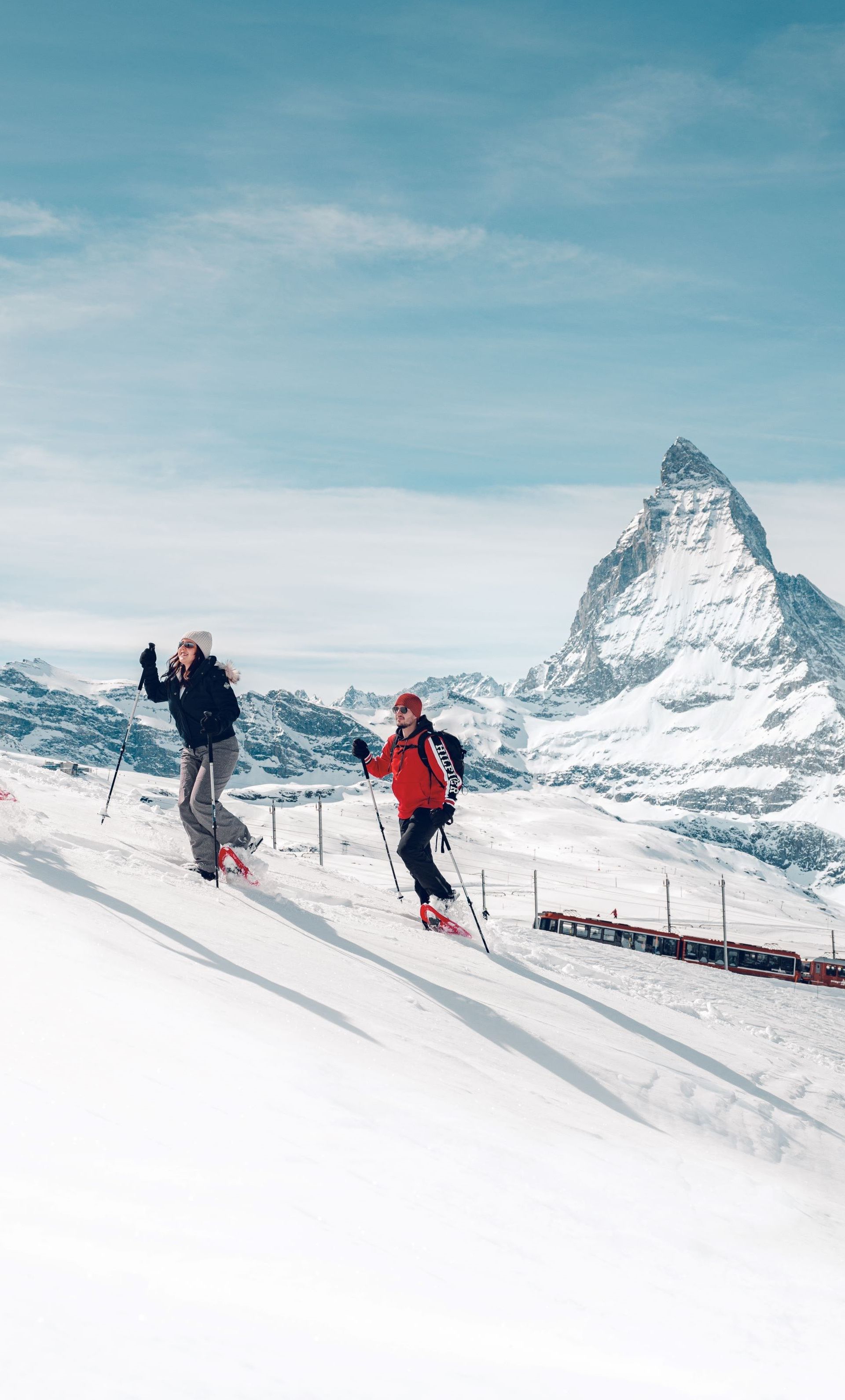 Drei Personen wandern mit Schneeschuhen durch eine verschneite Landschaft, im Hintergrund die Gornergrat Bahn und das Matterhorn.