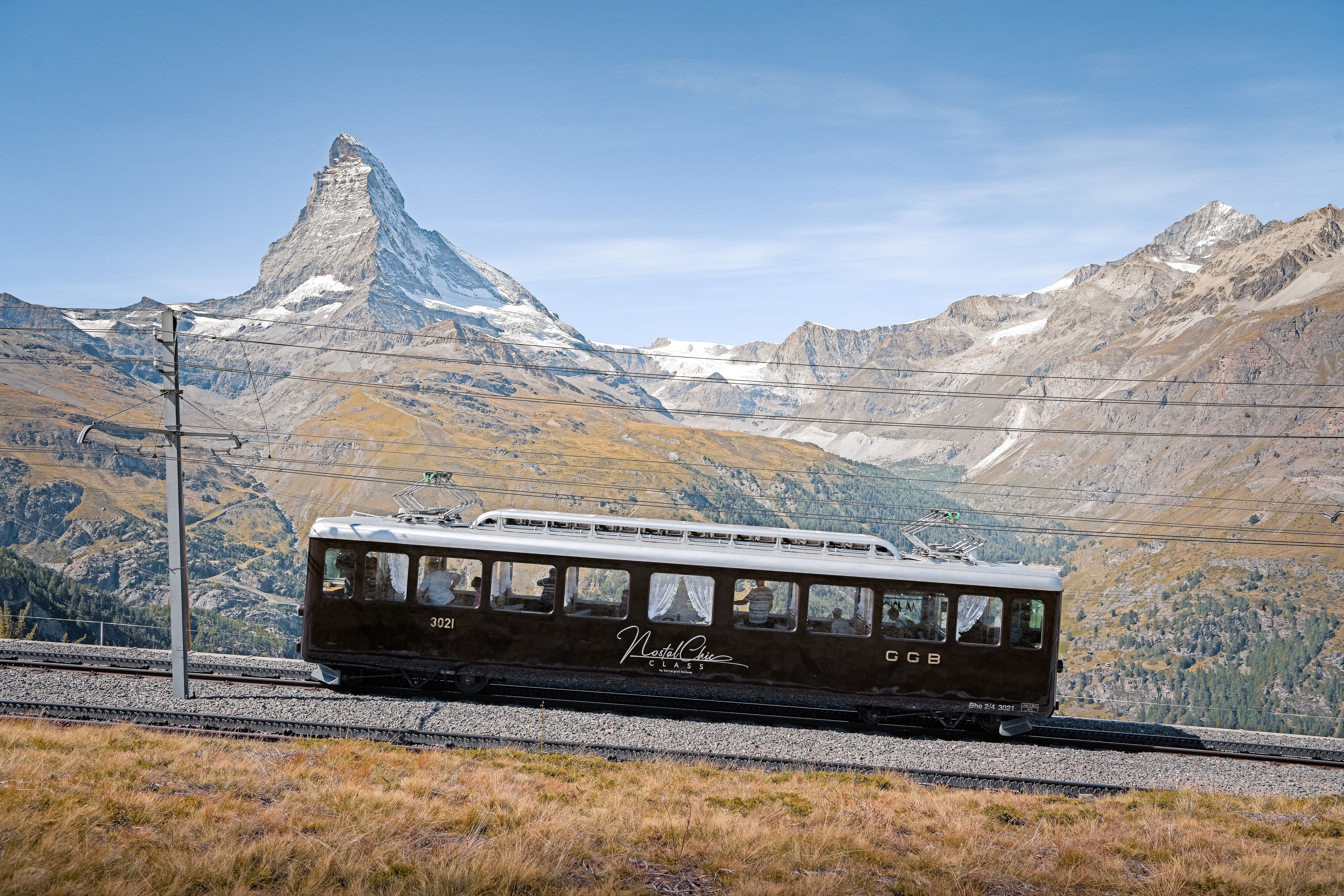 NostalChic Class Wagen auf der Strecke, Blick auf Matterhorn, Sommer Der braune NostalChic Class-Waggon fährt bei blauem Himmel auf das schneebedeckte Matterhorn zu. Die Landschaft in Braun- und Grüntönen fängt die sommerliche Stimmung ein.