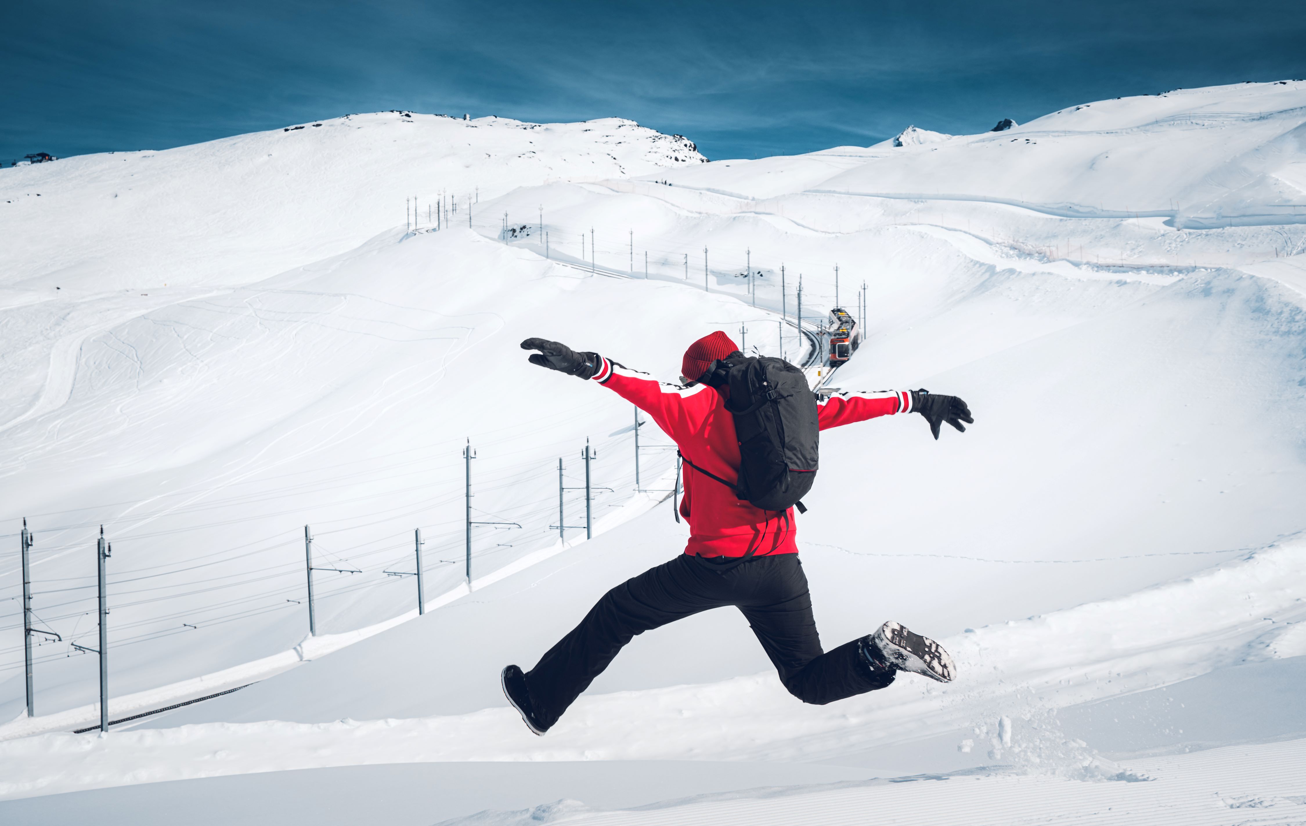 Ein Mann springt im Vordergrund, während im Hintergrund die Gonergrad Bahn und eine verschneite Landschaft zu sehen sind.